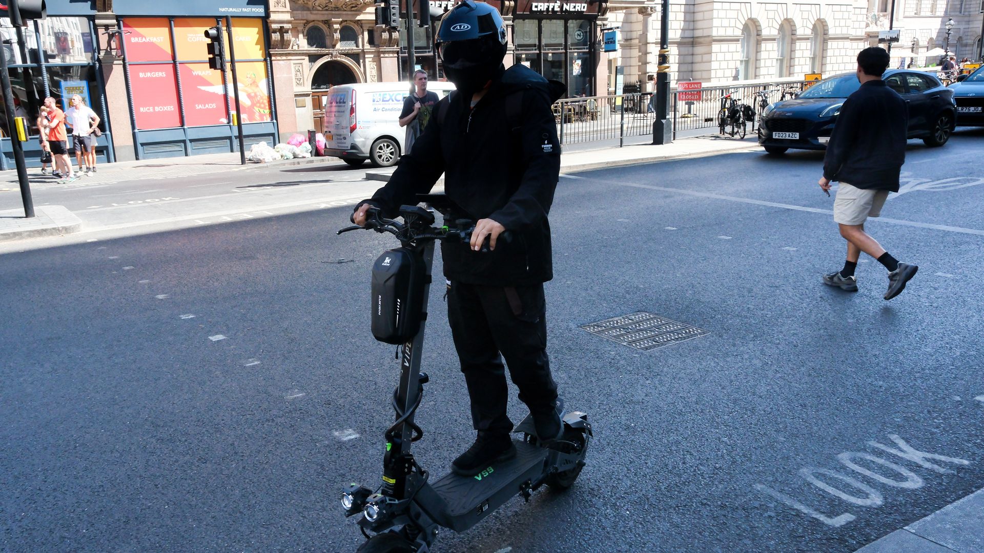 Person wearing black protective gear and a helmet rides a black electric scooter on a busy city street, with pedestrians, a white van, and a cafe storefront visible in the background.