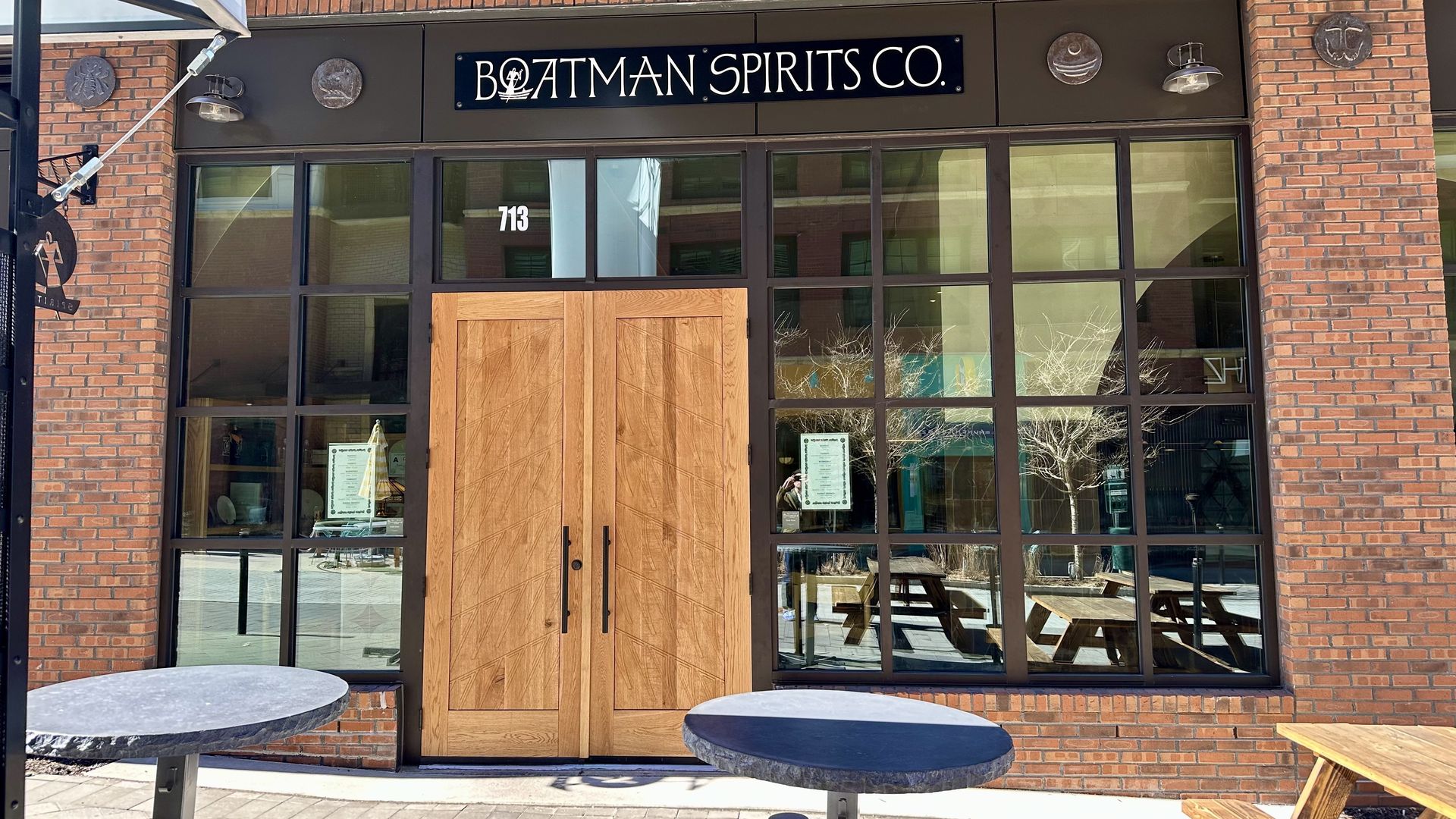 Brick building with large windows and wooden double doors; sign above reads BOATMAN SPIRITS CO.; outdoor tables and benches in front on a paved surface in daylight.