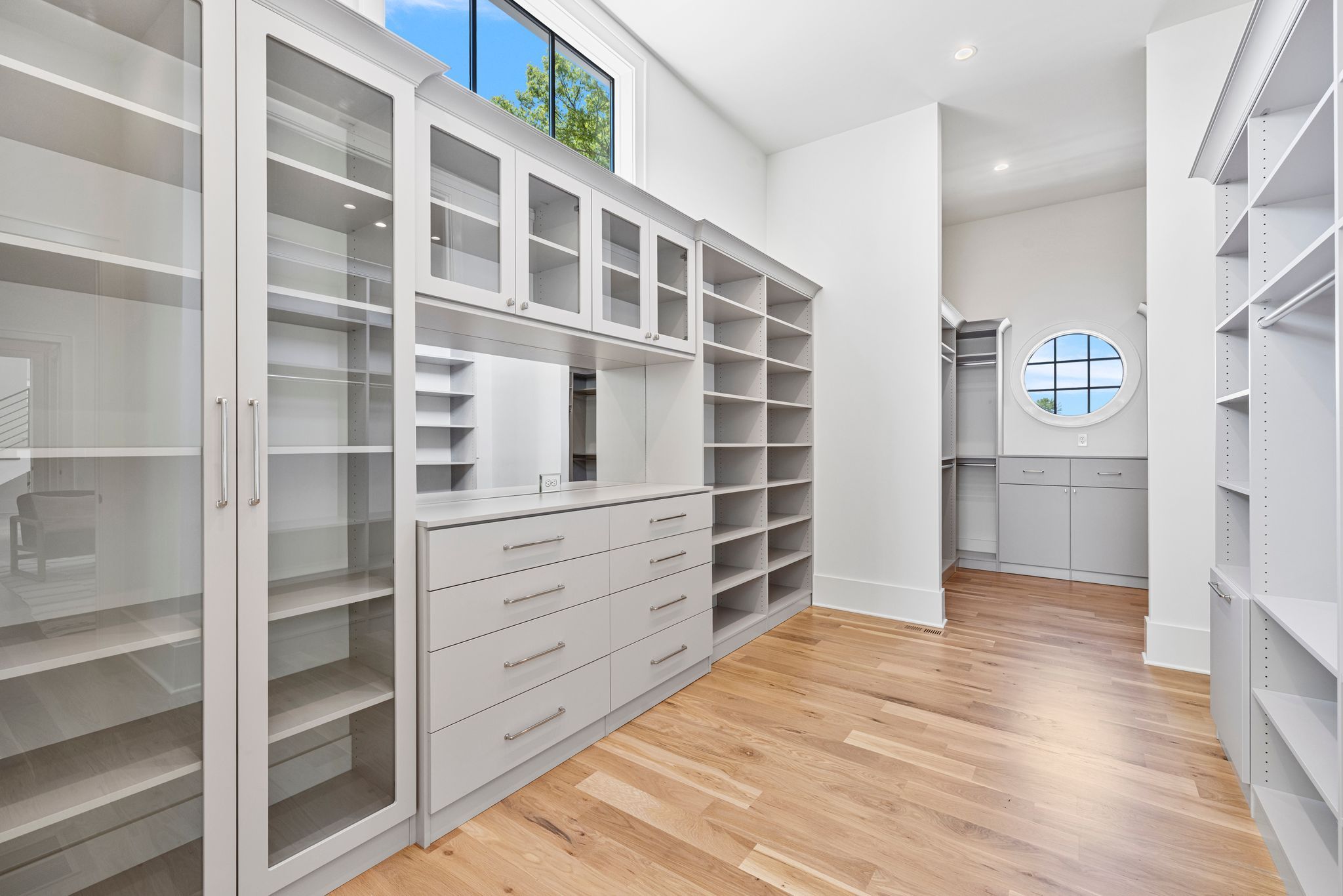 Spacious walk-in closet with light gray shelves and drawers, glass cabinet doors, a round window, and light wood flooring under soft ceiling lights.