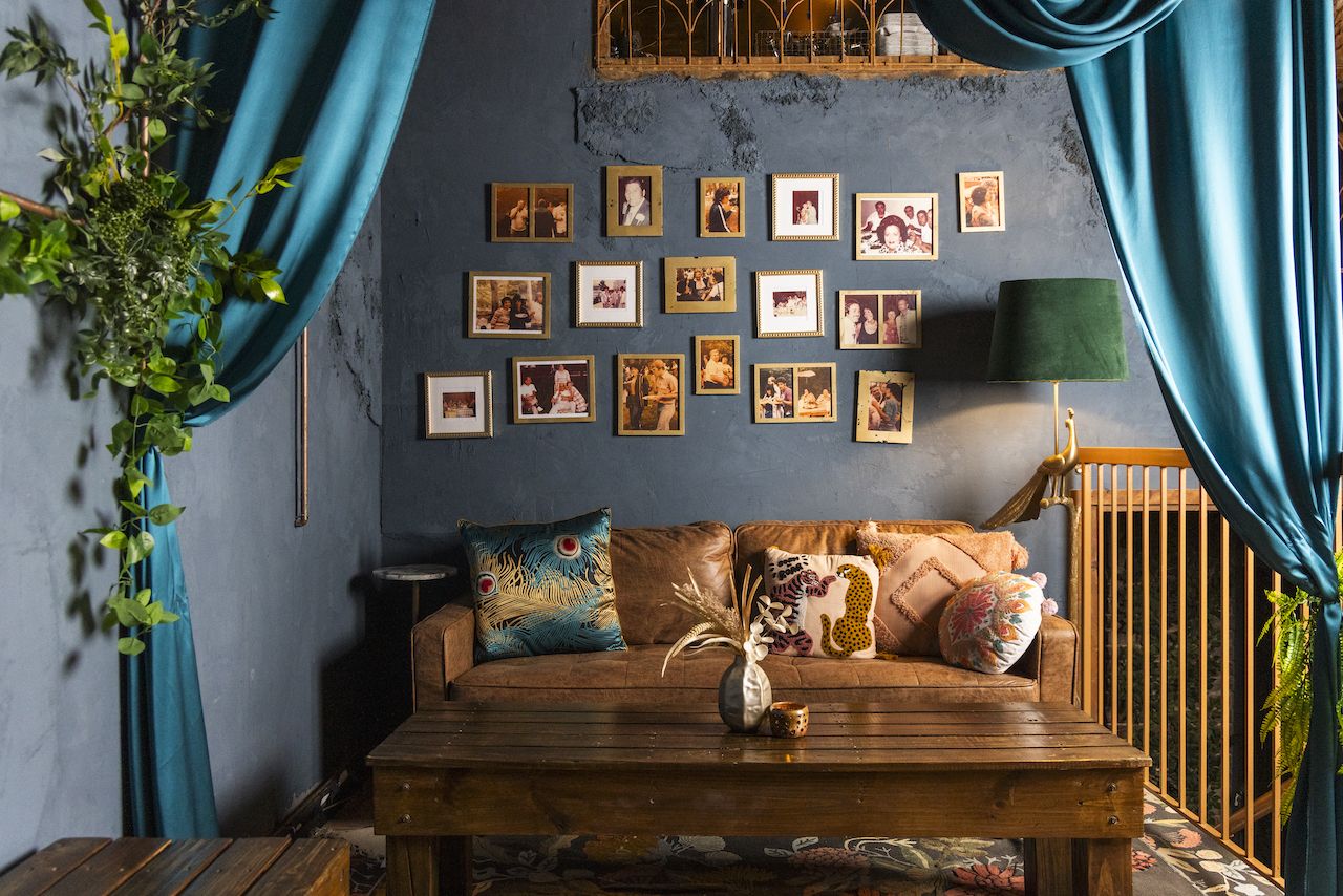 Cozy living room with brown leather sofa adorned with patterned pillows, dark teal curtains, green lamp, wooden coffee table, and a wall of various family photos in gold frames.