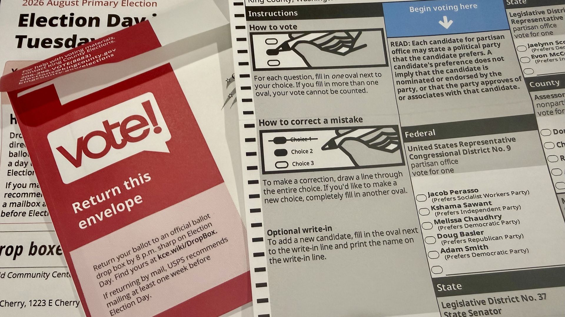 Close-up of election materials: a red Vote! envelope atop a white ballot stating Election Day Tuesday, plus a black-and-white instructions page with a blue Begin voting here banner.