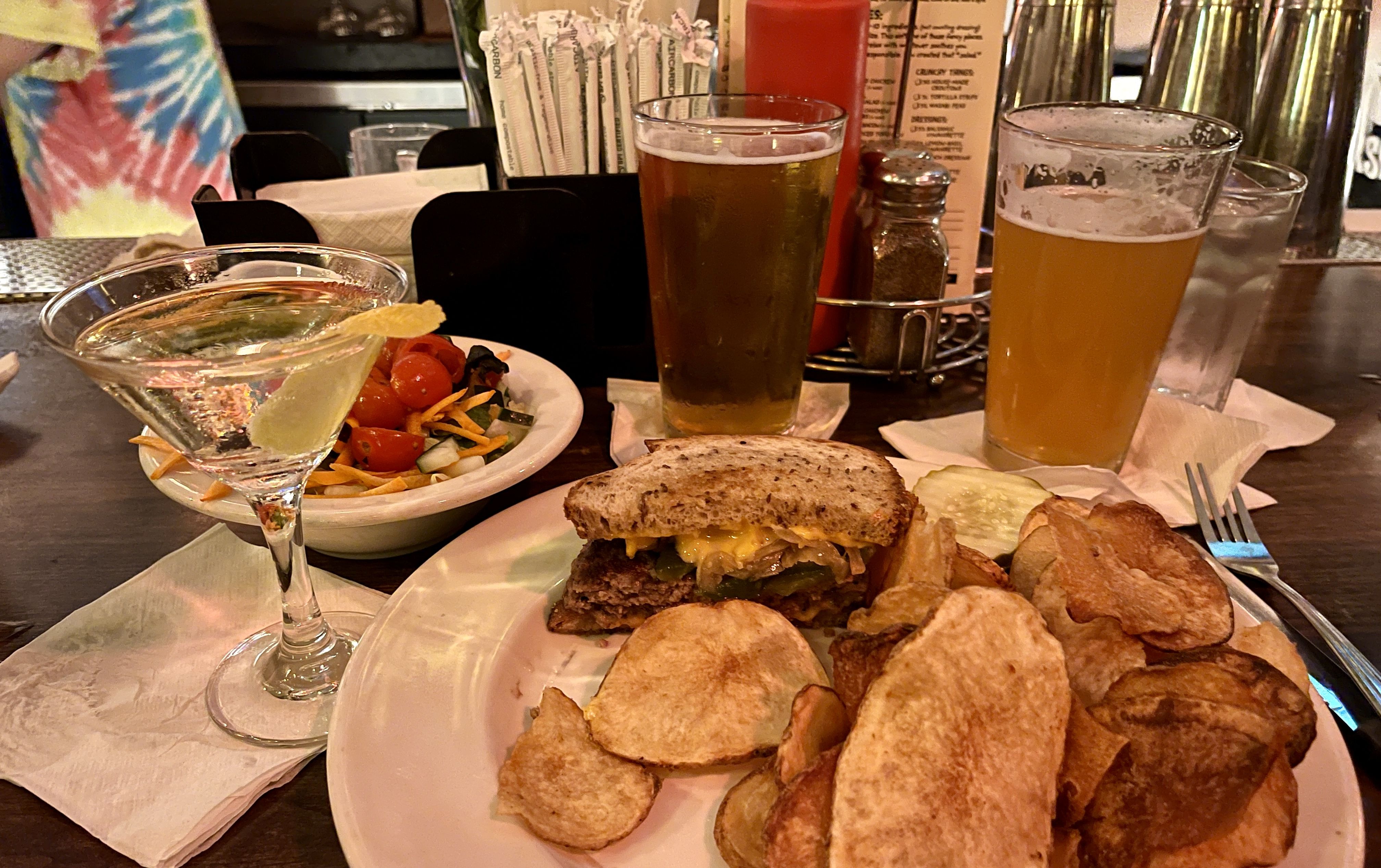 A meal at a bar with half a cheeseburger, homemade potato chips, a dill pickle slice, side salad with tomatoes and carrots, a martini with lemon twist, a glass of beer, and a hazy beer.