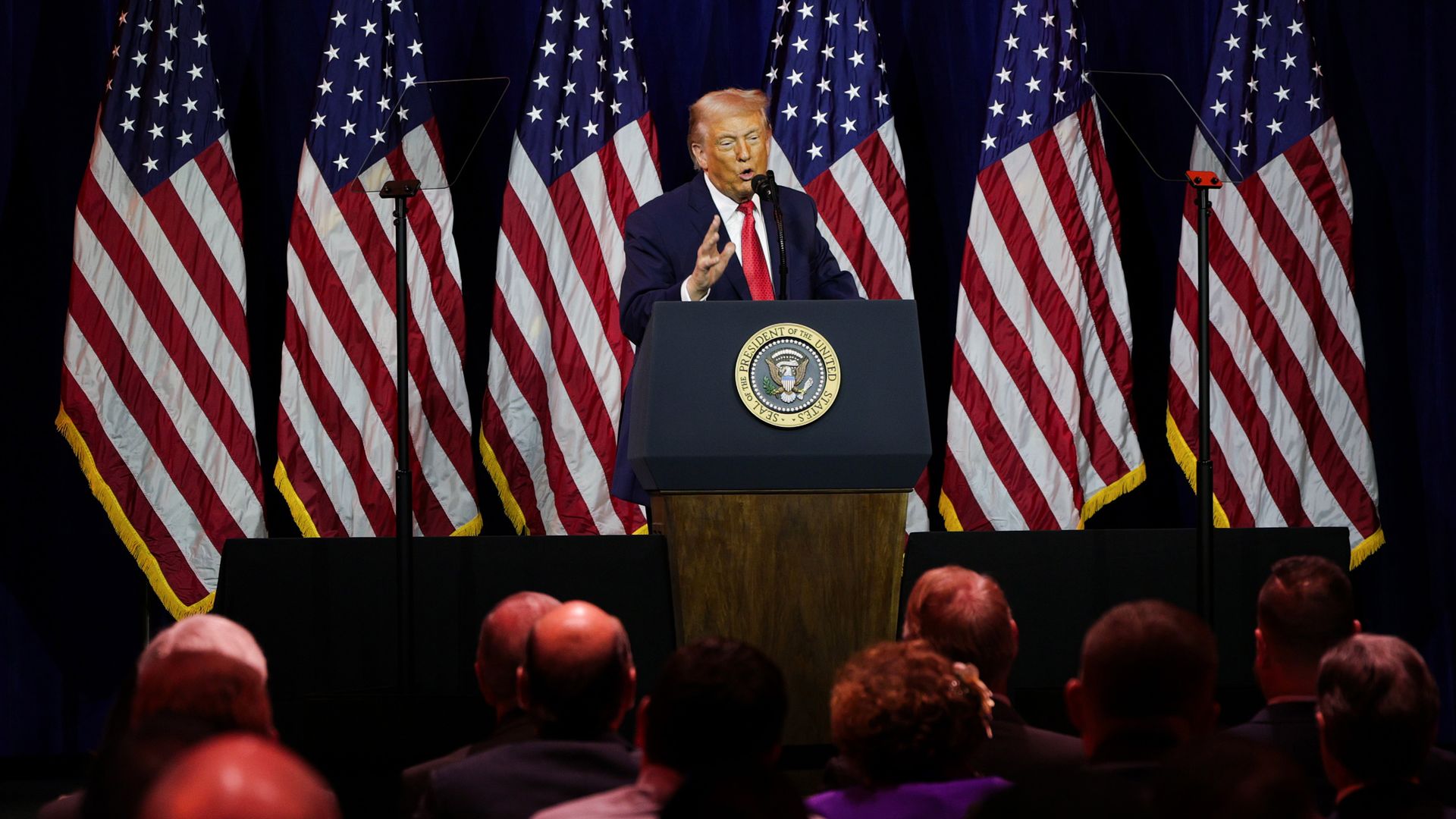 A man in a navy suit with a red tie speaks at a podium with the U.S. presidential seal, in front of multiple American flags, addressing an audience seated in a darkened room.