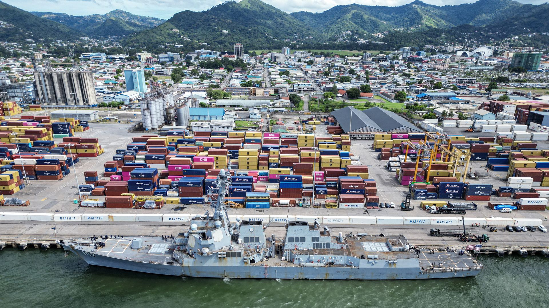 Aerial view of the USS Gravely warship docked in the port of Port of Spain, Trinidad and Tobago, on October 26, 2025. Brightly colored shipping containers can be seen at the doc in front of buildings, with green hills in the background.