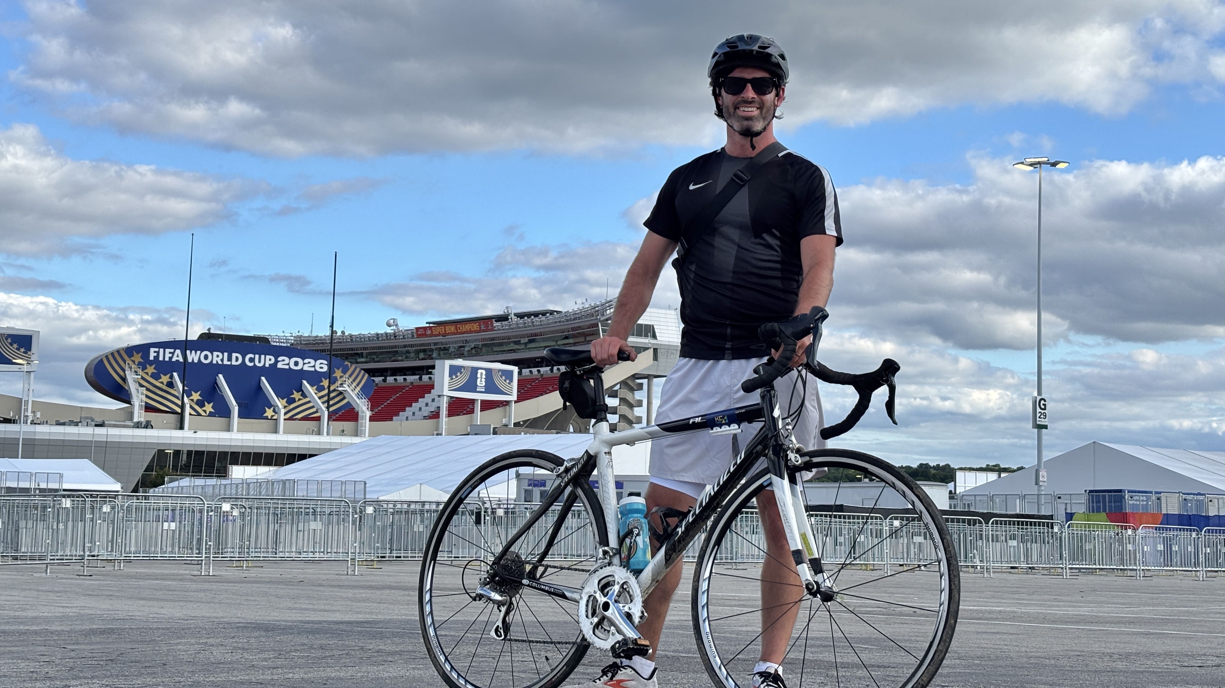 Man wearing black and gray cycling kit, helmet, and sunglasses stands with a road bike at an outdoor stadium. A large "FIFA World Cup 2026" banner and a blue sky with clouds behind.