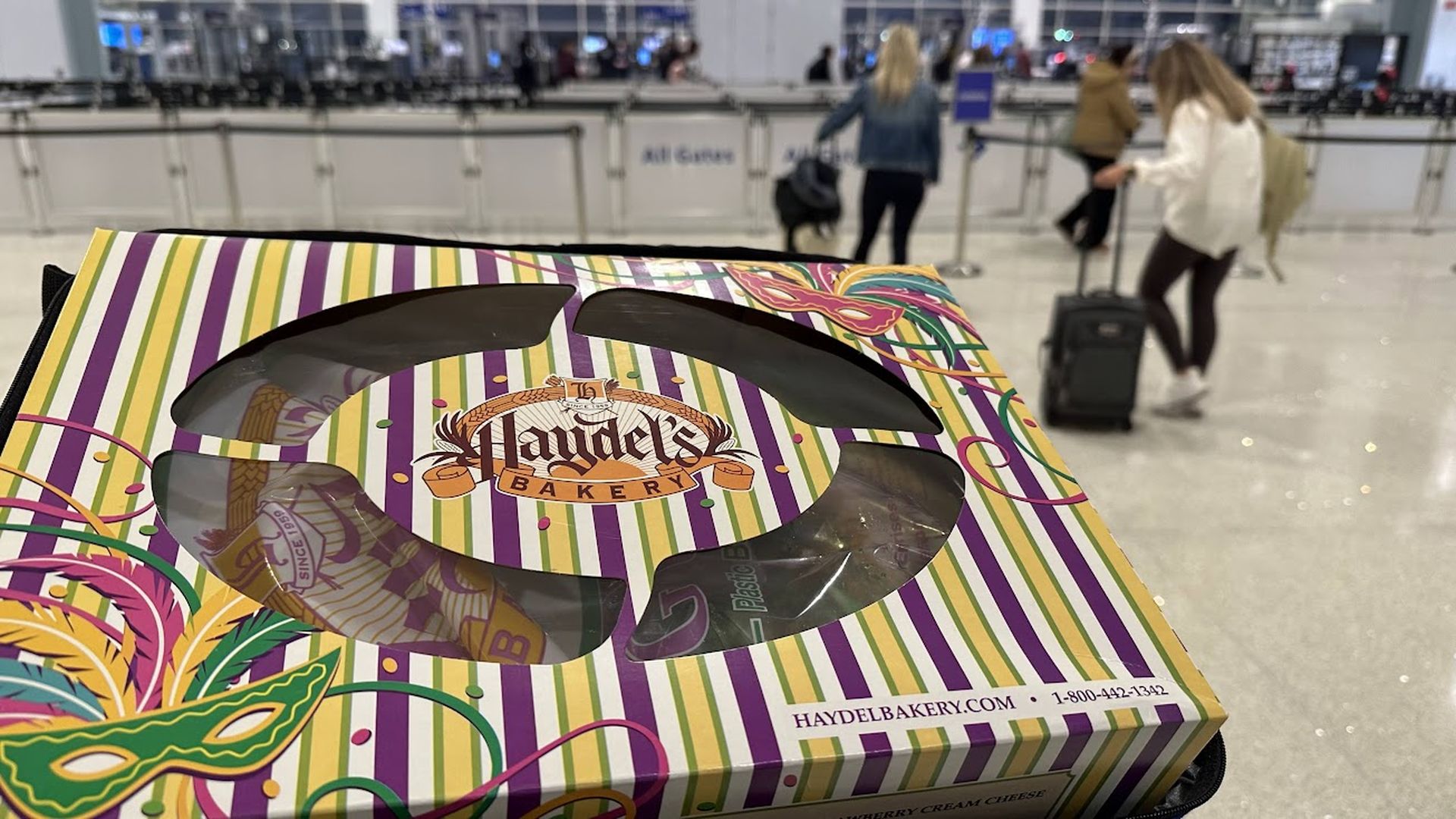 Colorful striped Haydel's Bakery box with Mardi Gras mask design held in an airport with blurred travelers and check-in counters in the background.