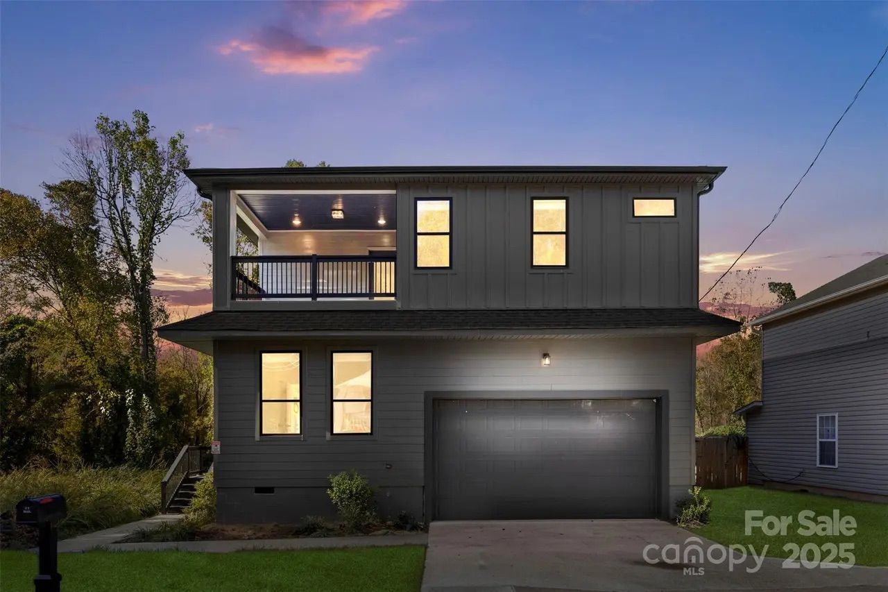 Two-story gray house with garage and lit windows at dusk, partly cloudy sky with pink and purple hues, green grass in front, and neighboring houses visible.