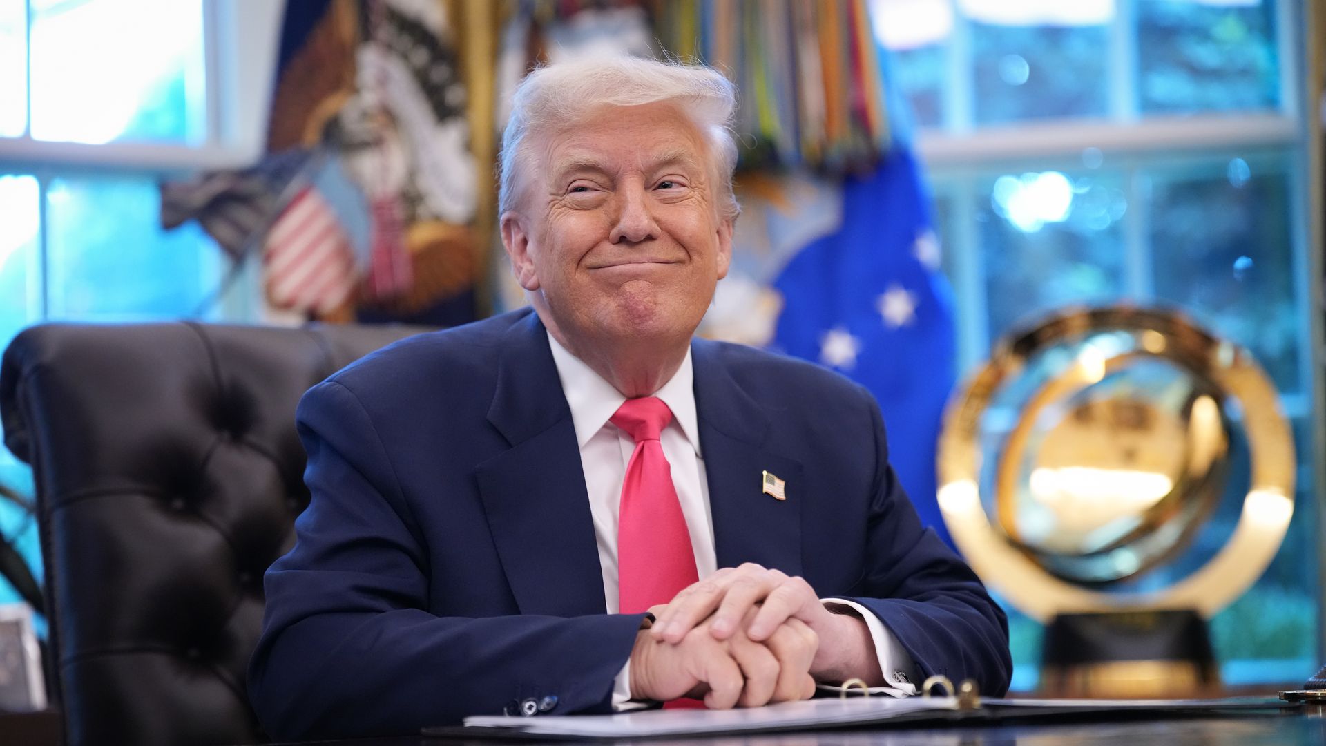 President Trump, wearing a navy suit and red tie, sitting at a desk with hands clasped, smiling slightly in an office with flags and a golden globe in the background.