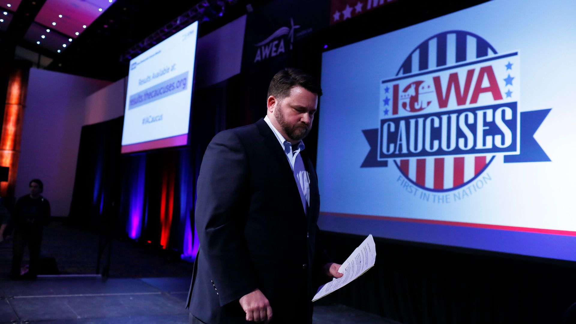 In this image, a man in a suit walks across a stage while holding paper. A screen behind him says "Iowa Caucuses" 