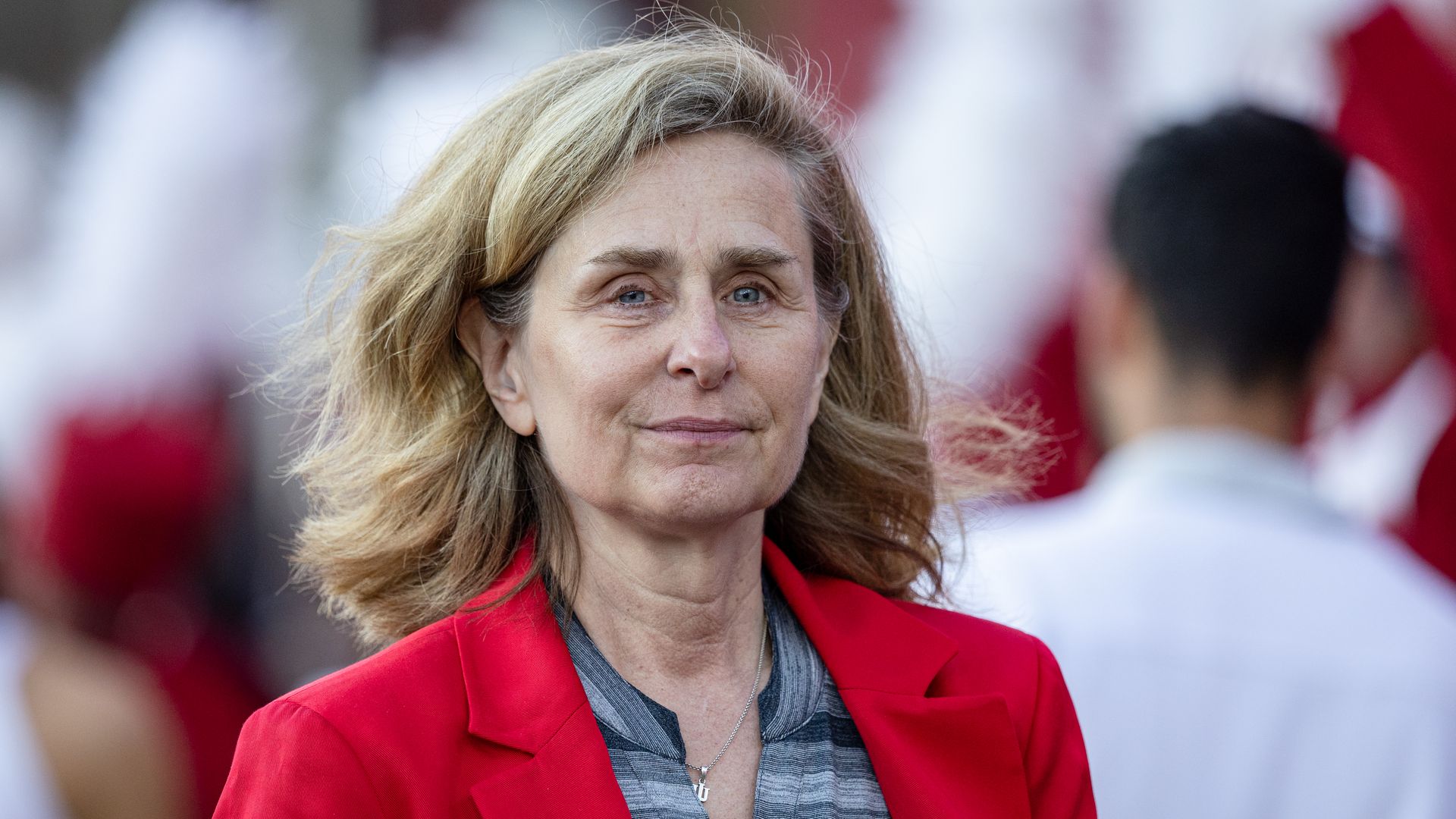 Woman with blonde hair wearing a red jacket and gray striped shirt with "Indiana" logo, outdoors with blurred people and red-white colors in the background.