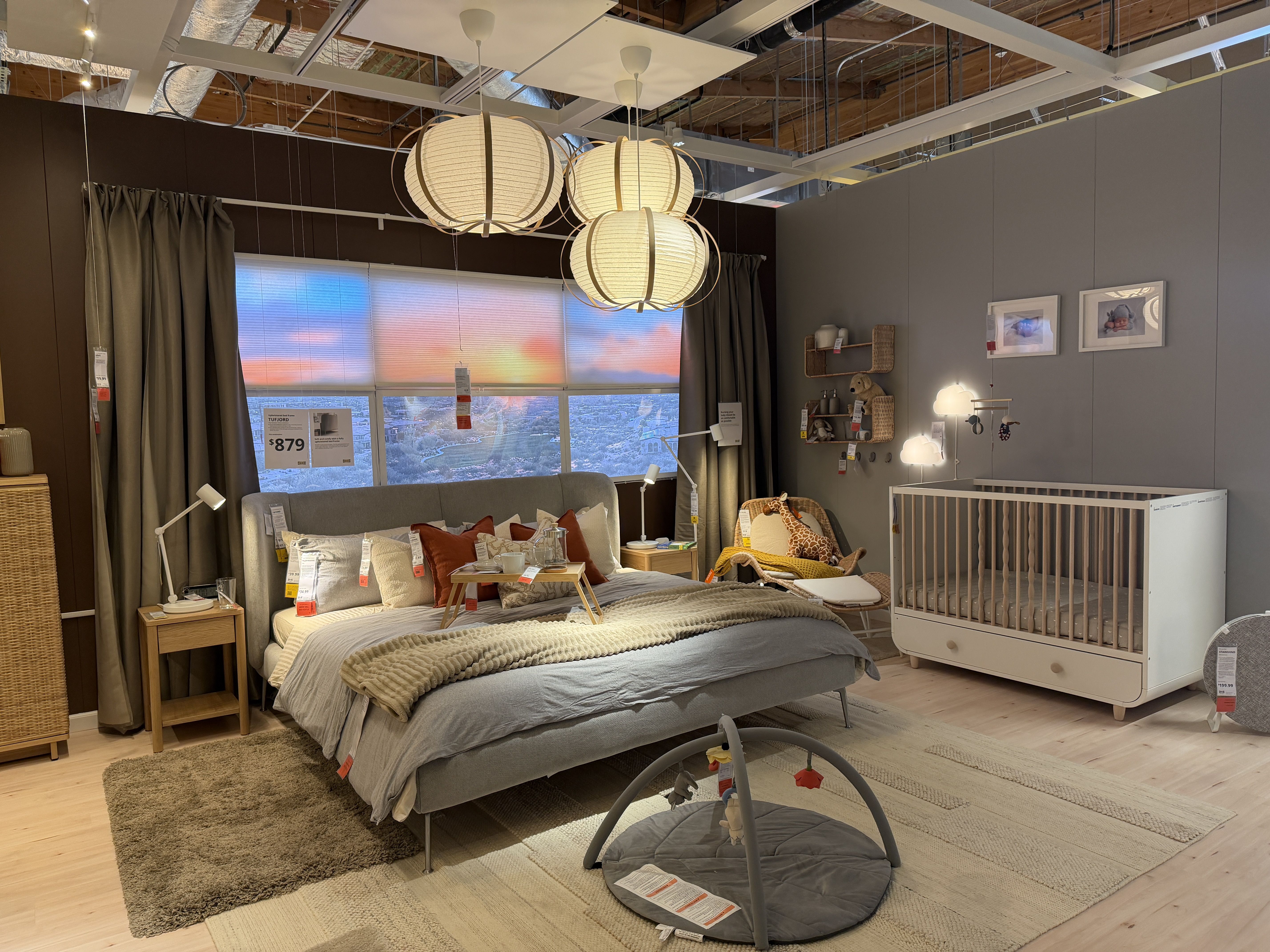 Cozy showroom bedroom with a gray upholstered bed, beige throws, and red/cream pillows. Nearby crib, wicker chair, wooden nightstands, and hanging paper lanterns; blinds glow through window.