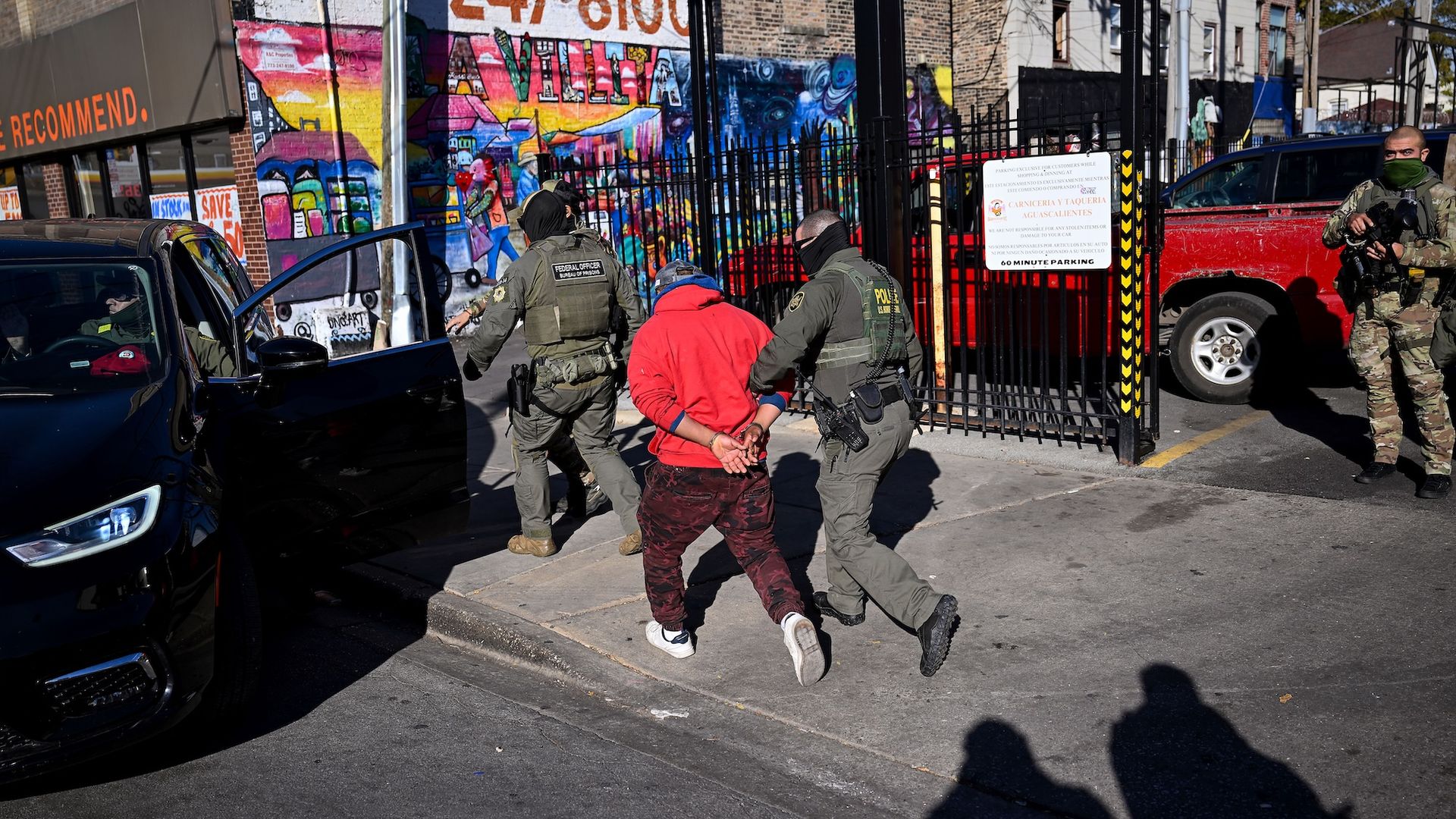 A U.S. Customs and Border Patrol agent holding the arm of a man in a red sweatshirt whose hands are tied behind his back walking behind another agent getting into a black car.