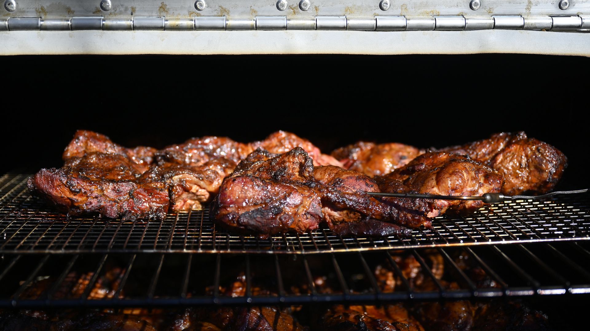 Close-up of several pieces of well-seasoned, grilled meat cooking on metal racks inside a smoker or grill, glowing with rich browns and charred textures.