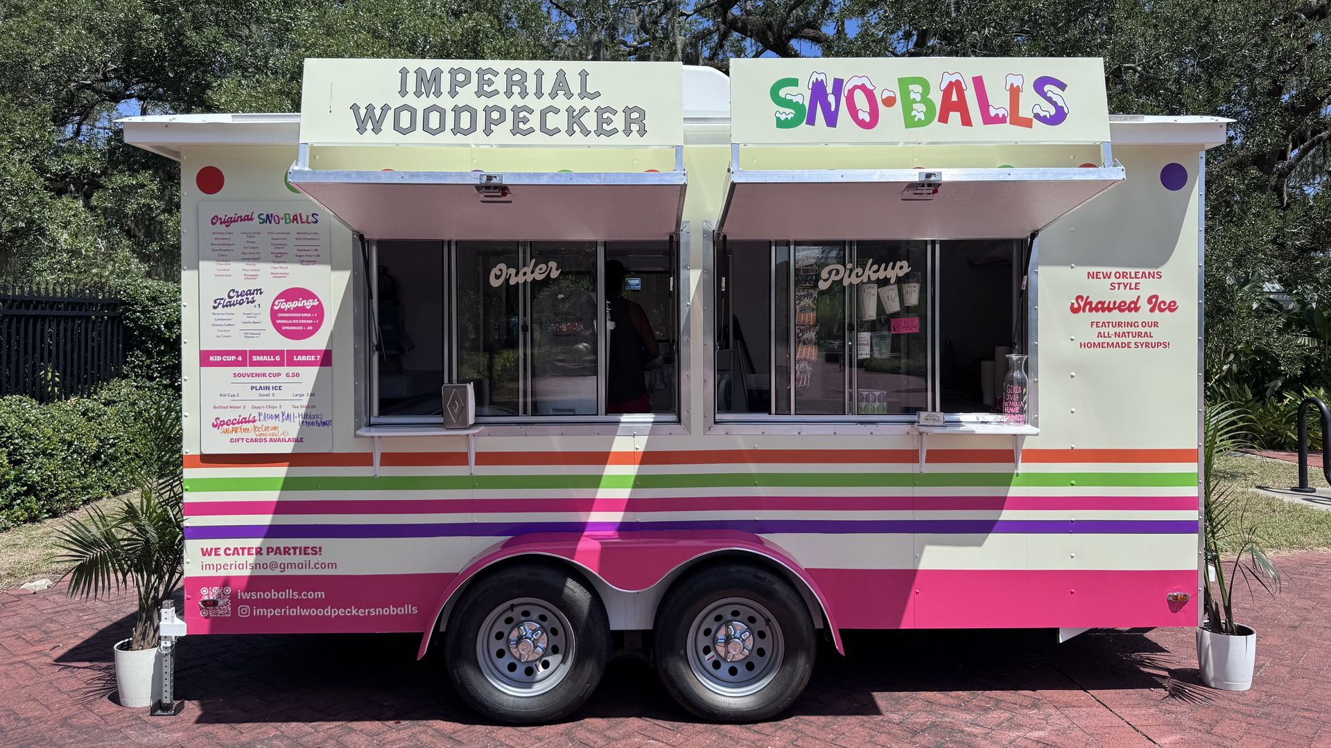Colorful food truck with signs "Imperial Woodpecker" and "Sno-Balls"; pastel yellow body, pink and green stripes, open windows, and a backdrop of trees under a bright blue sky.