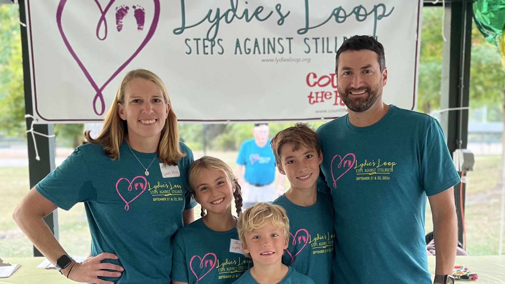 A smiling family of two adults and three children wearing matching teal T-shirts with pink heart logos stand in front of a banner that reads "Lydie's Loop Steps Against Stillbirth."