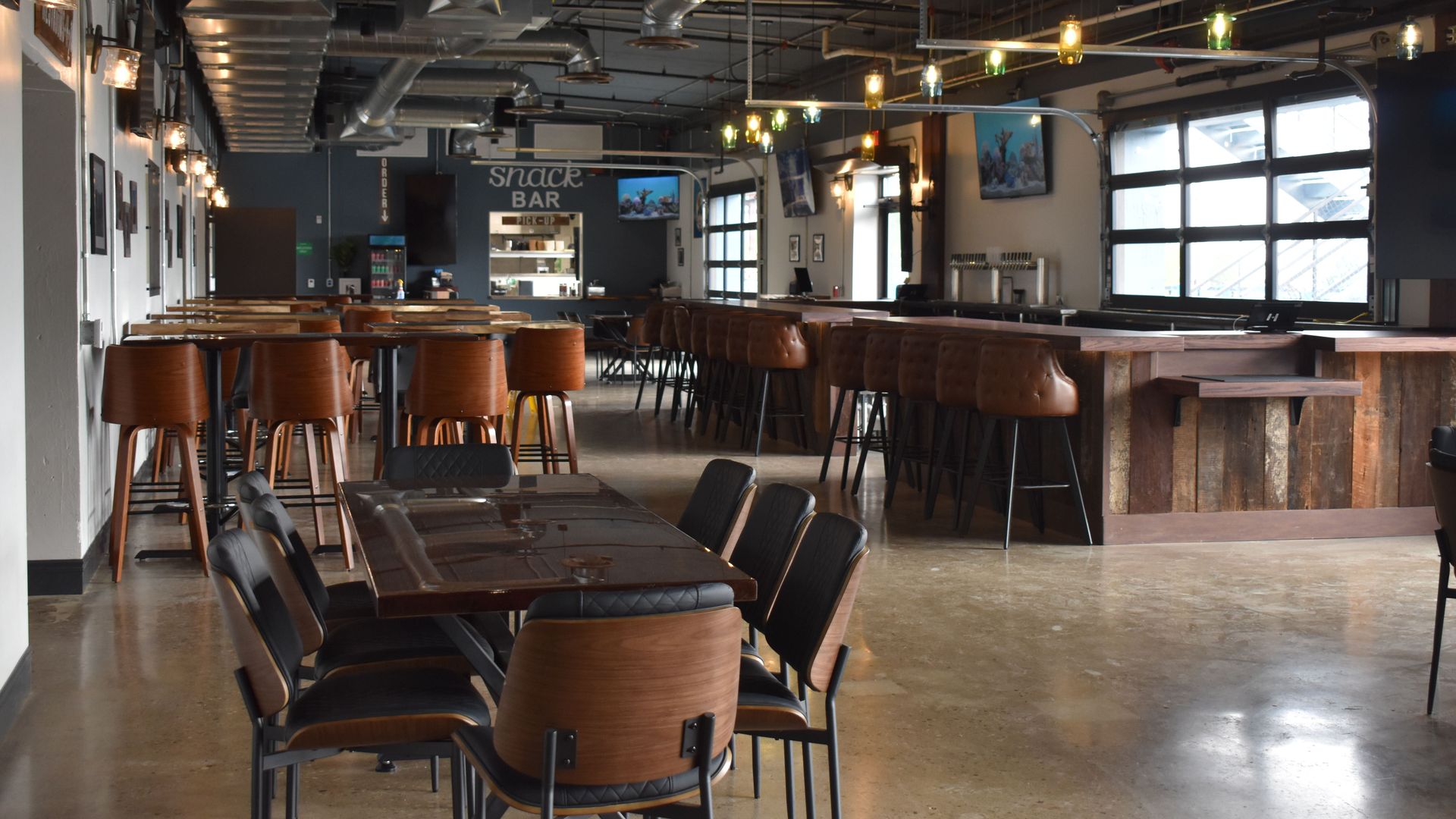 Empty modern bar and snack area with wooden tables, brown leather and black chairs, industrial ceiling with ducts, and windows letting natural light inside.