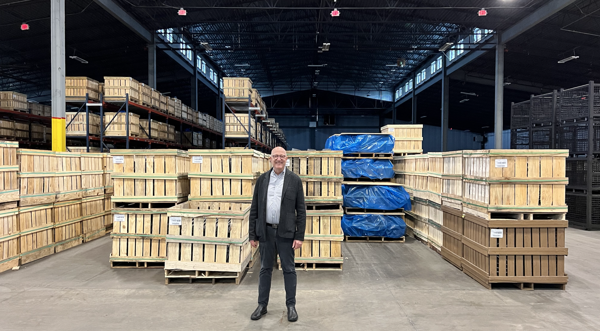 man stands in front of industrial warehouse with boxes and crates of materials behind him