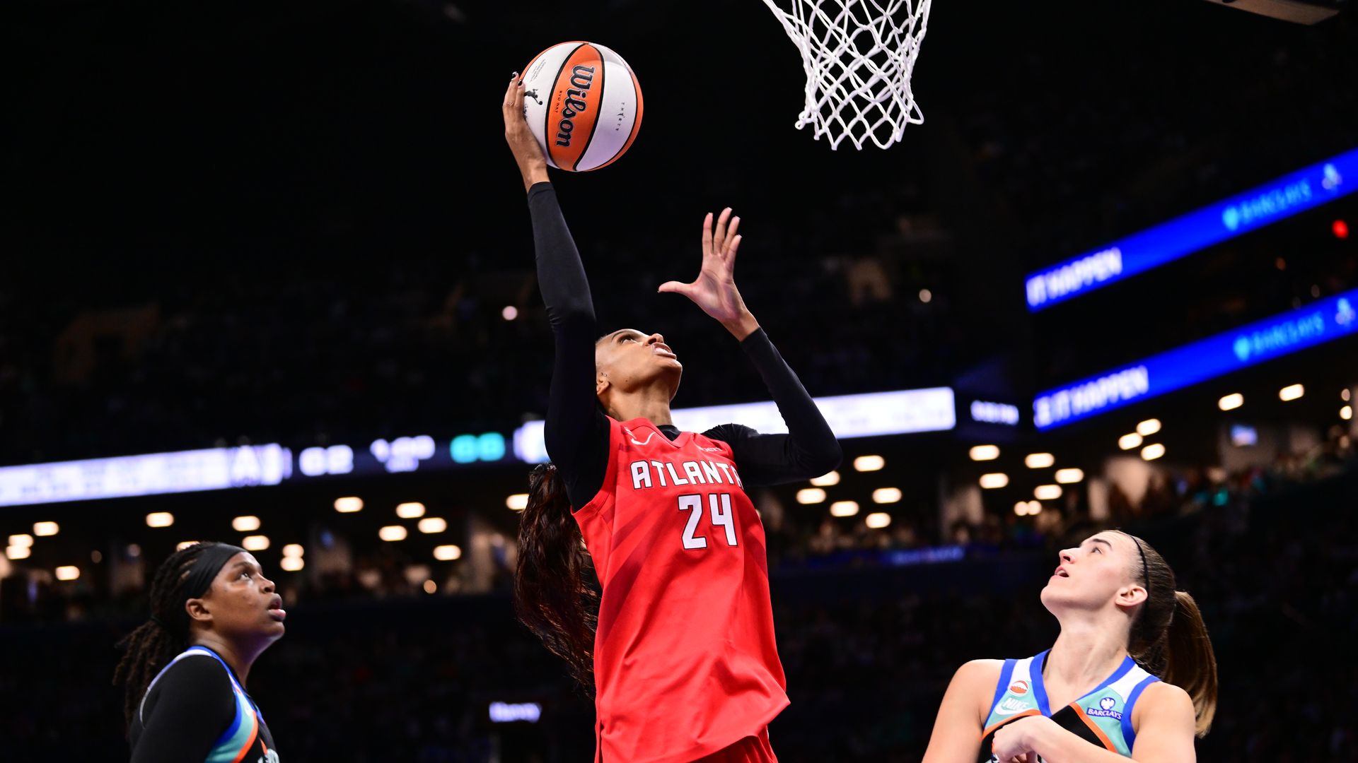 A female athlete wearing a red jersey with the word ATLANTA and number 24 written on front, red shorts and black compression sleeves holds a basketball in her hand as she tries to drive to the basket. Two players wearing black, blue, white and green jerseys stand on the left and right.