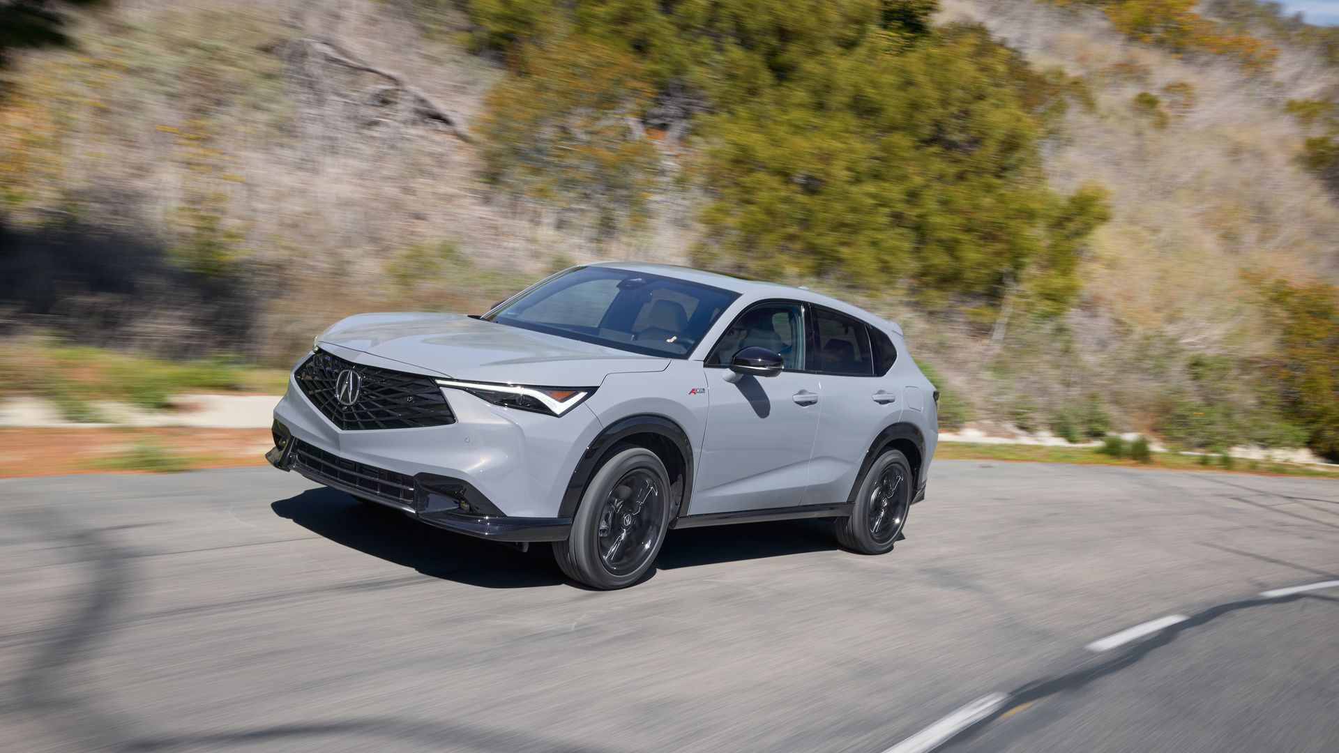 Gray Acura ADX SUV driving on a winding road with blurred background of green and leafless trees on a sunny day.