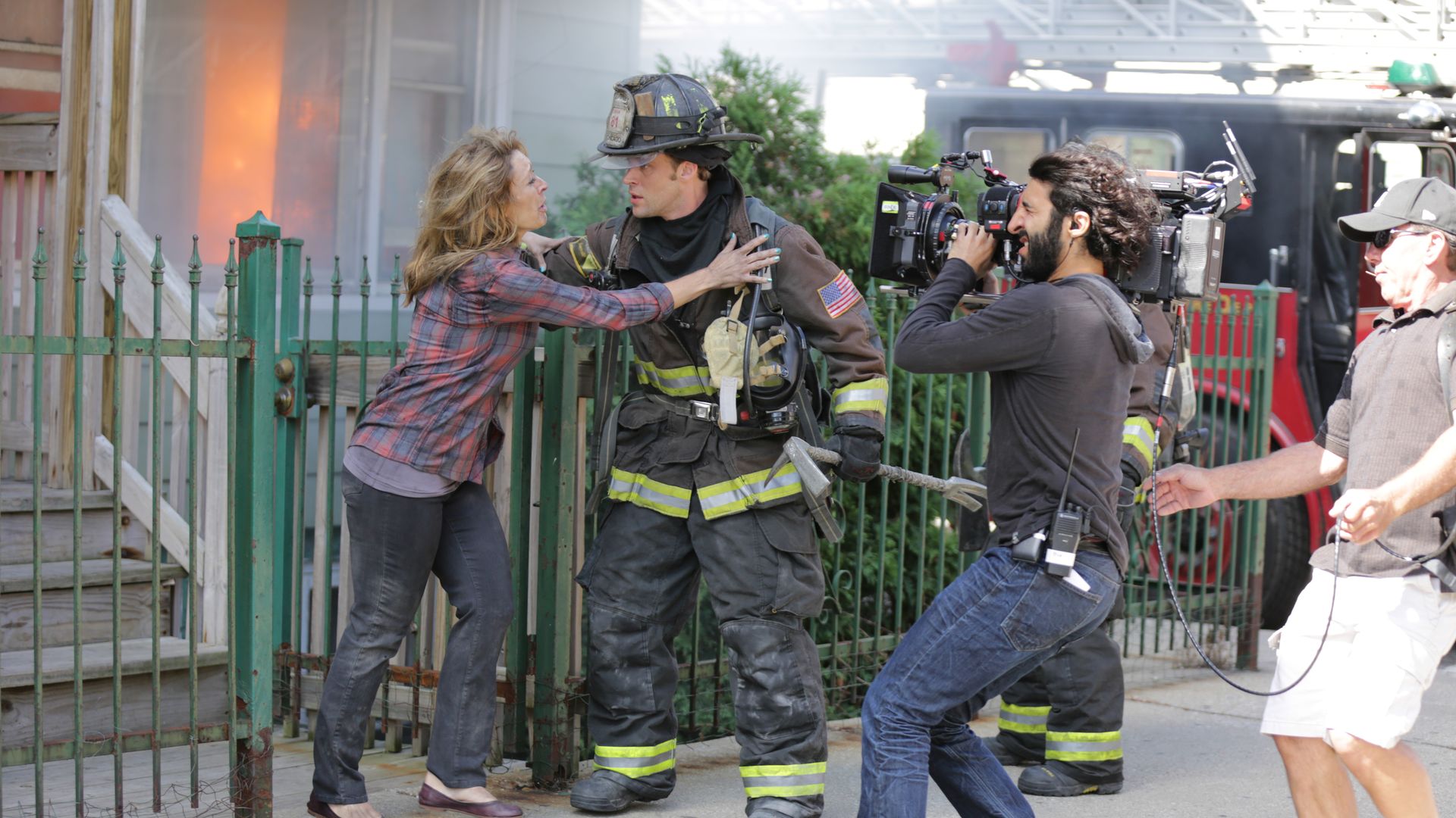 Female civilian in a plaid shirt argues with a firefighter in full gear while a film crew records; orange flames glow from a burning building behind them, with a fire truck and green fence nearby.
