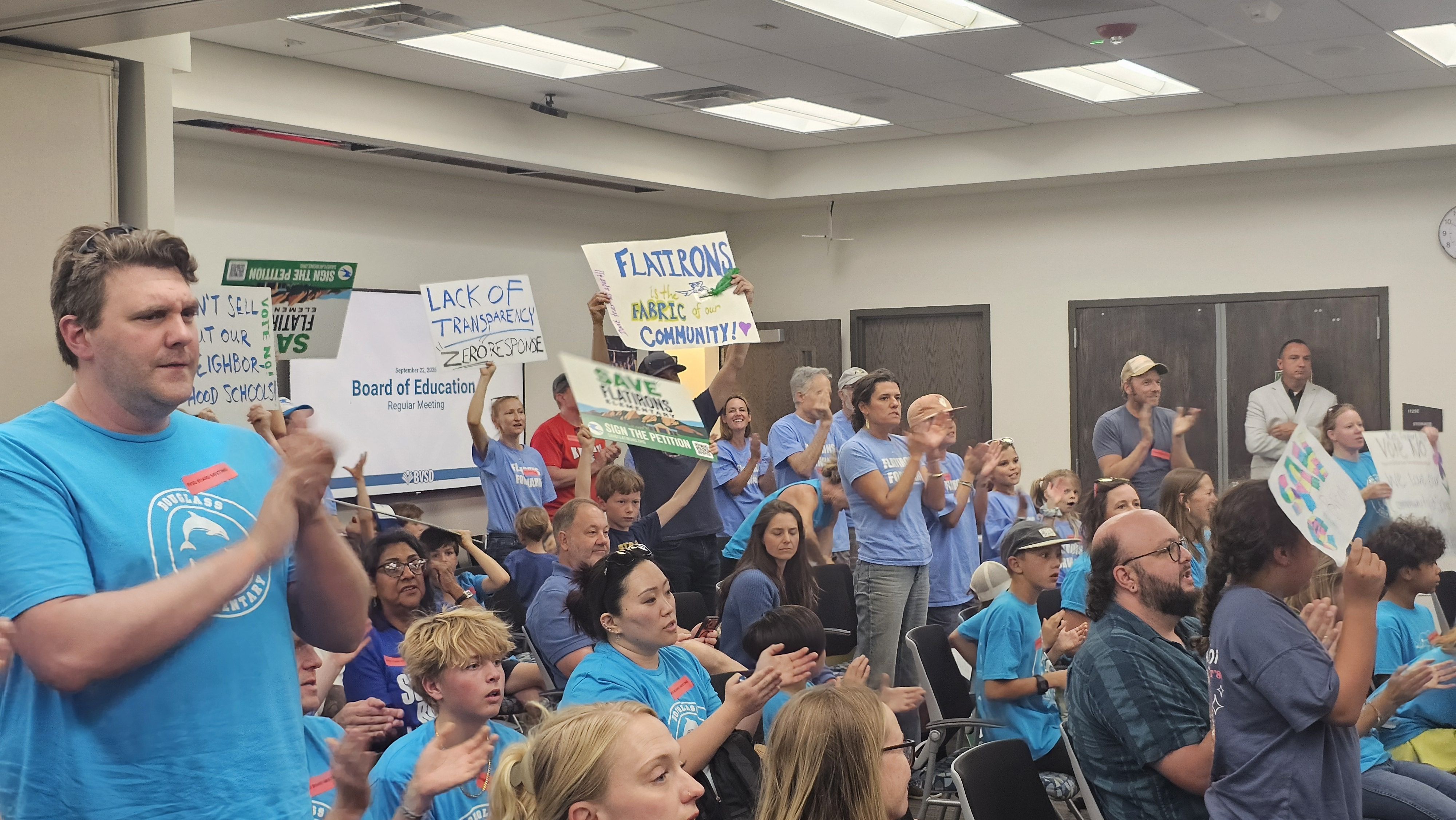 Crowd in a meeting room, many in blue shirts, holding signs and clapping. Signs protest lack of transparency and praise Flatirons as the community fabric.