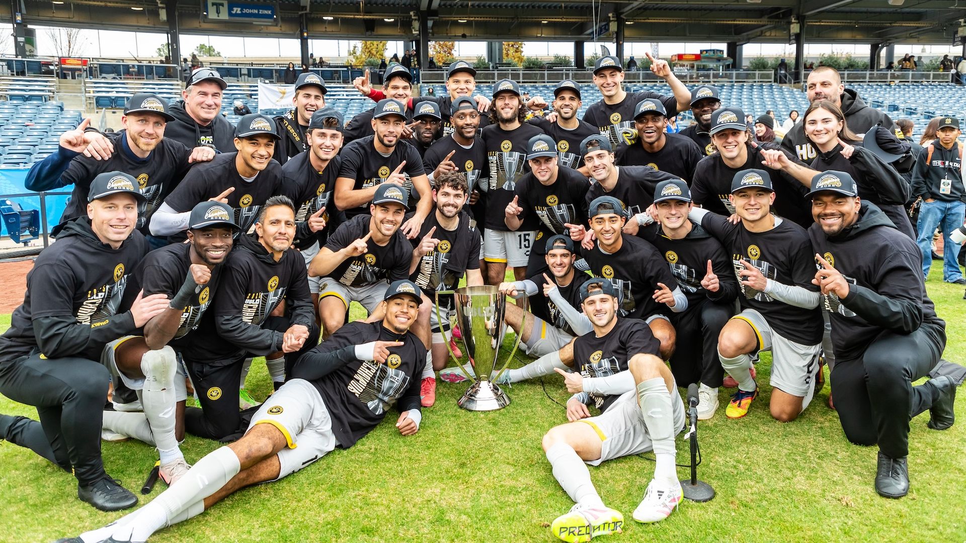 A soccer team wearing black championship hats and shirts poses on the field around a large trophy, all smiling and holding up number one fingers in celebration.