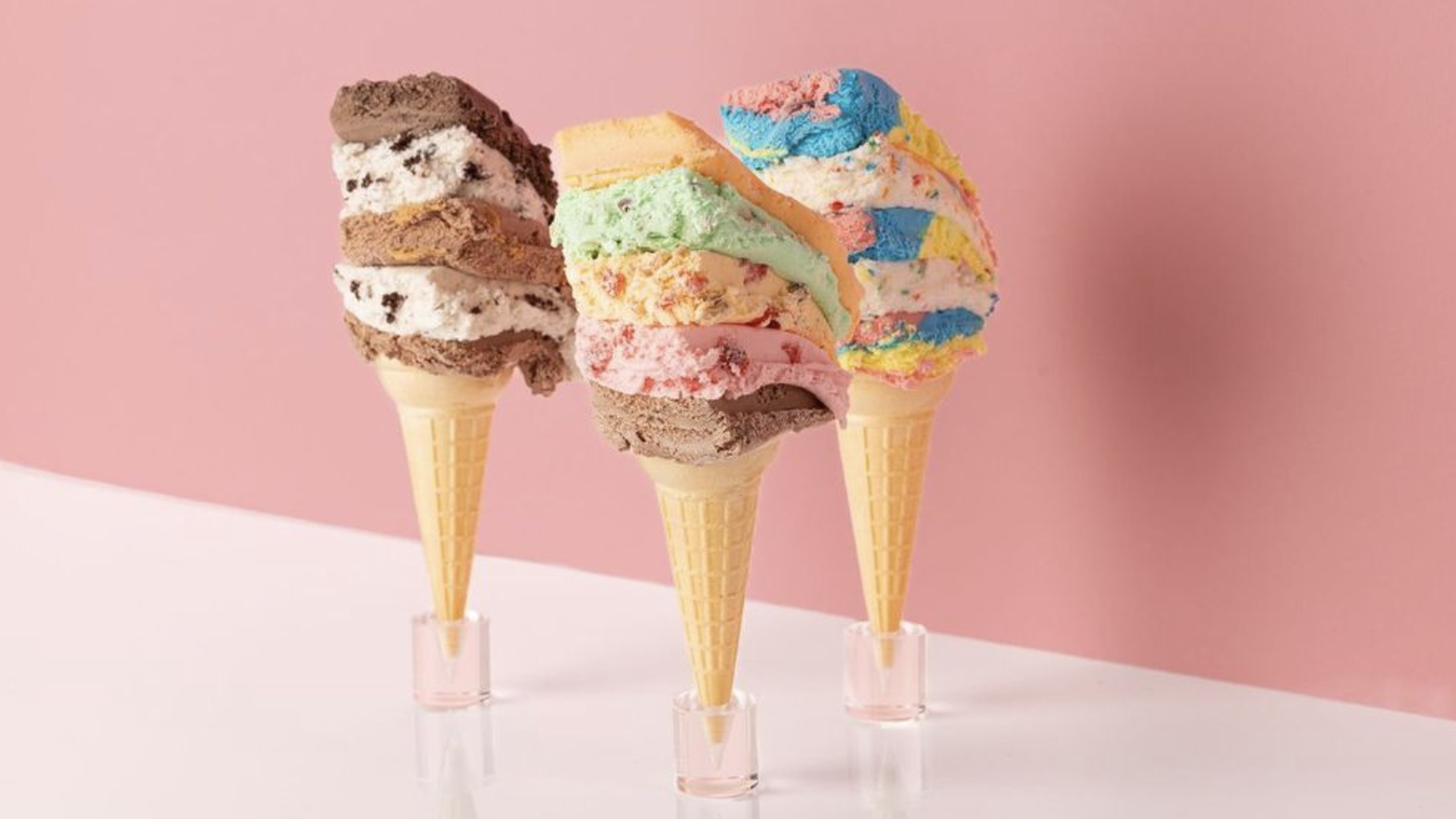Three ice cream cones with multicolored slices of chocolate, cookies and cream, mint, and rainbow ice cream stacked on them, on a pale pink backdrop; each cone rests in a small clear cup.