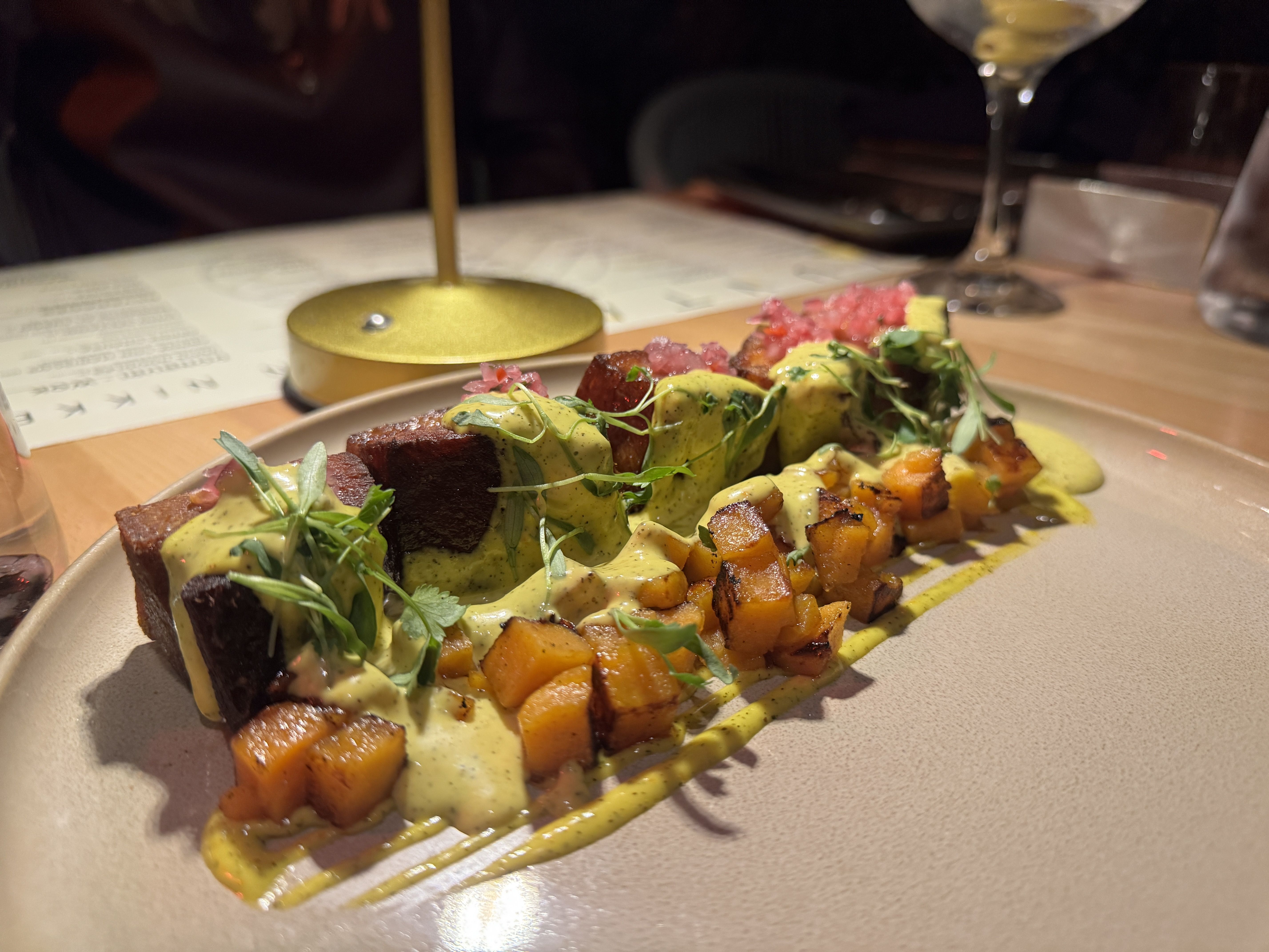 Plate of roasted potatoes, greens, and meat with a creamy yellow sauce, topped by pink edible flowers; blurred wine glass and restaurant table in the background.