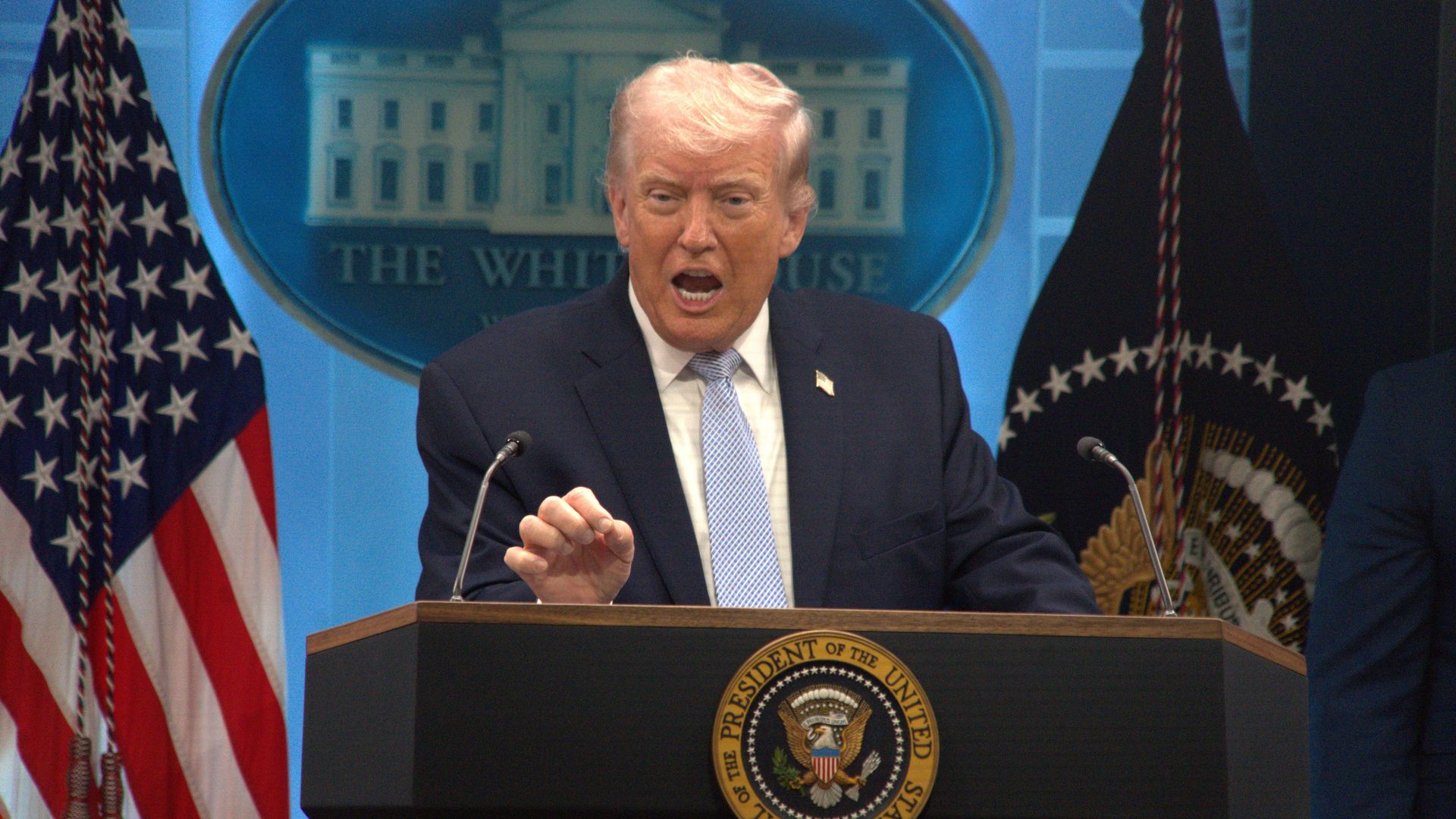 Man in a dark suit and light blue tie speaks at a podium with the presidential seal, flanked by American flags, against a blue backdrop featuring the White House.