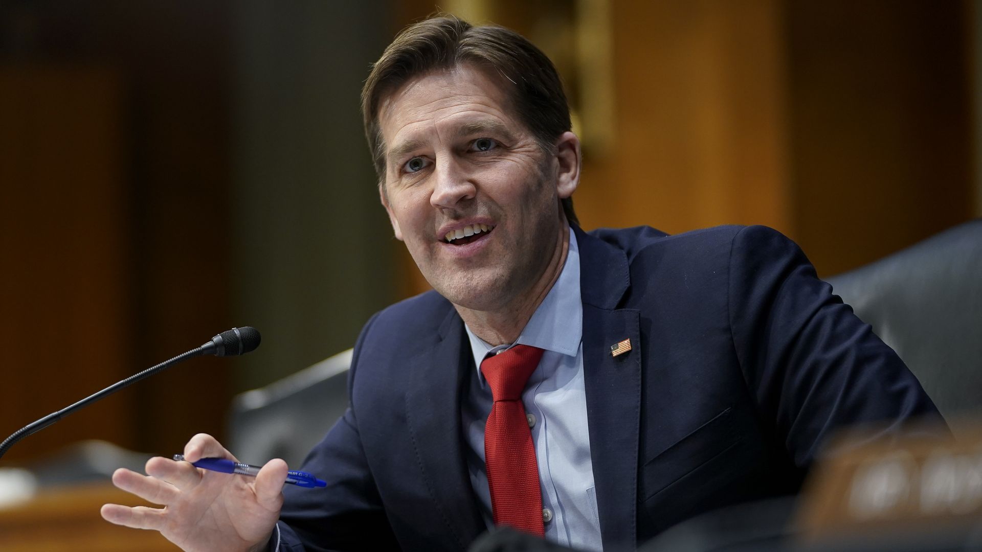 Smiling man in a navy suit and red tie, wearing an American flag pin, speaks into a microphone at a hearing, gesturing with a blue pen; wood-paneled backdrop.