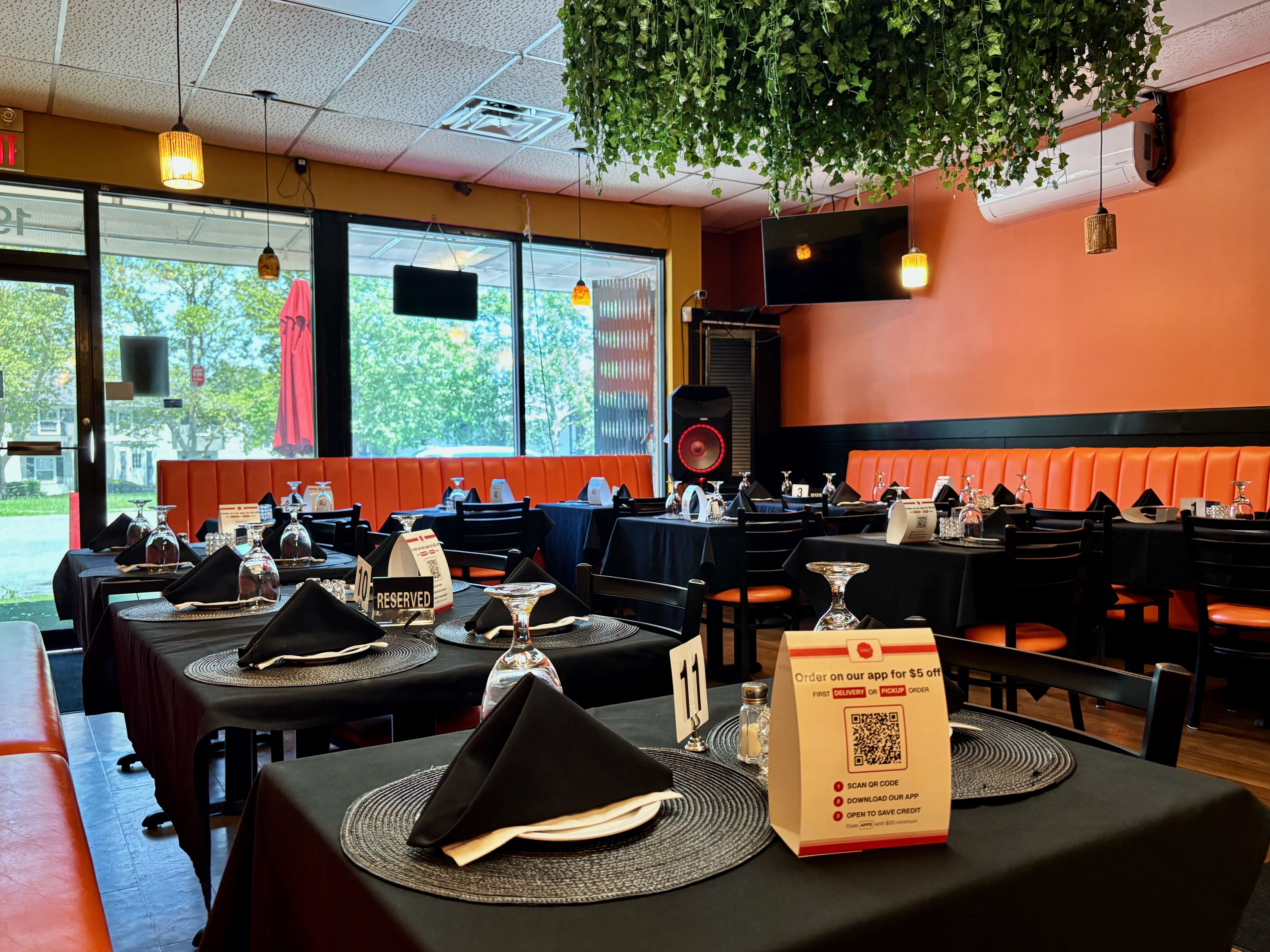 Interior of a restaurant with black tablecloths, black chairs, orange cushioned benches, hanging plants, and windows showing outdoor greenery on a sunny day.