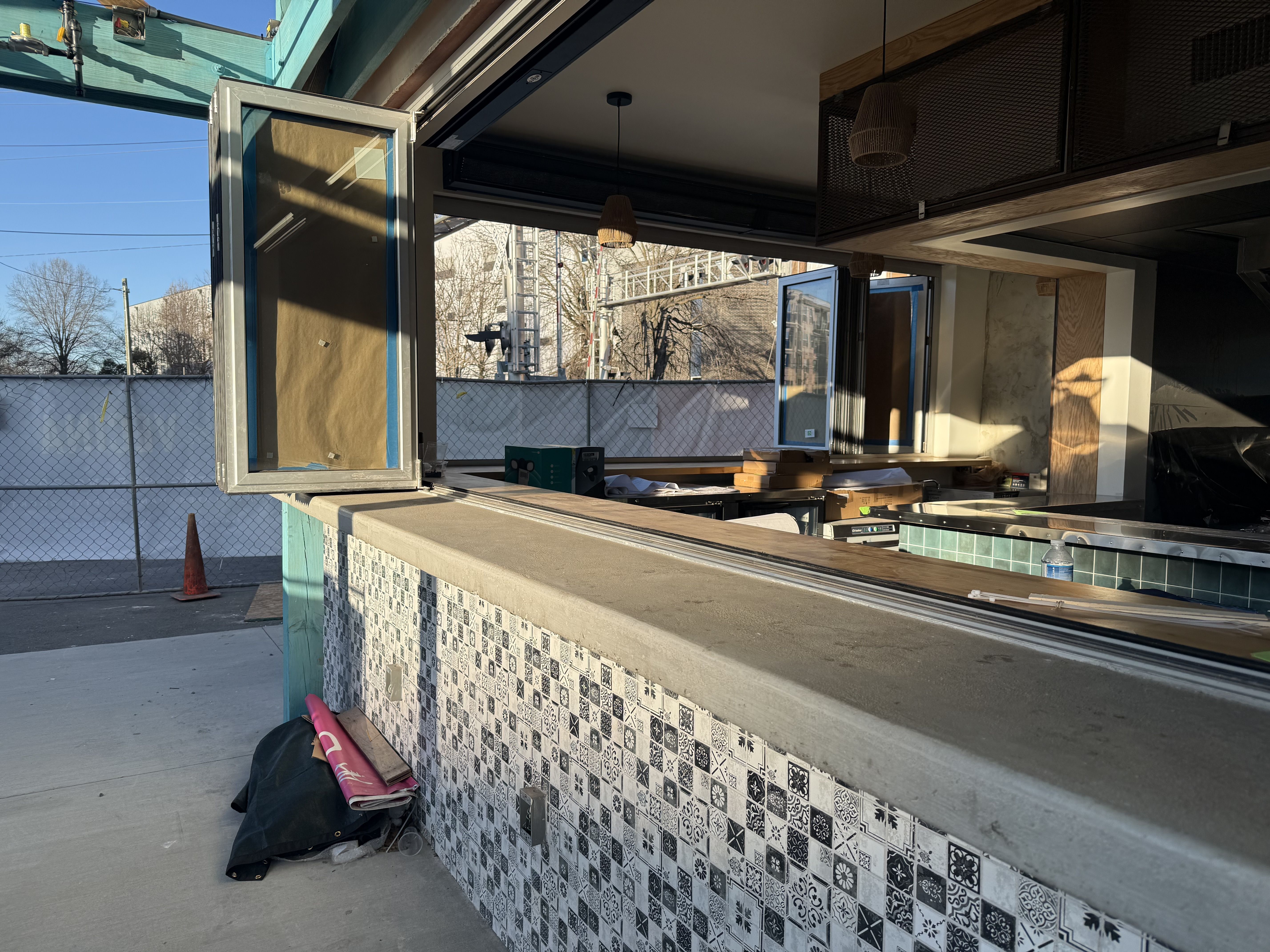 Outdoor view of a partially open food or drink service window with patterned black-and-white tiled counter and concrete top, under warm sunlight in a construction or renovation setting.