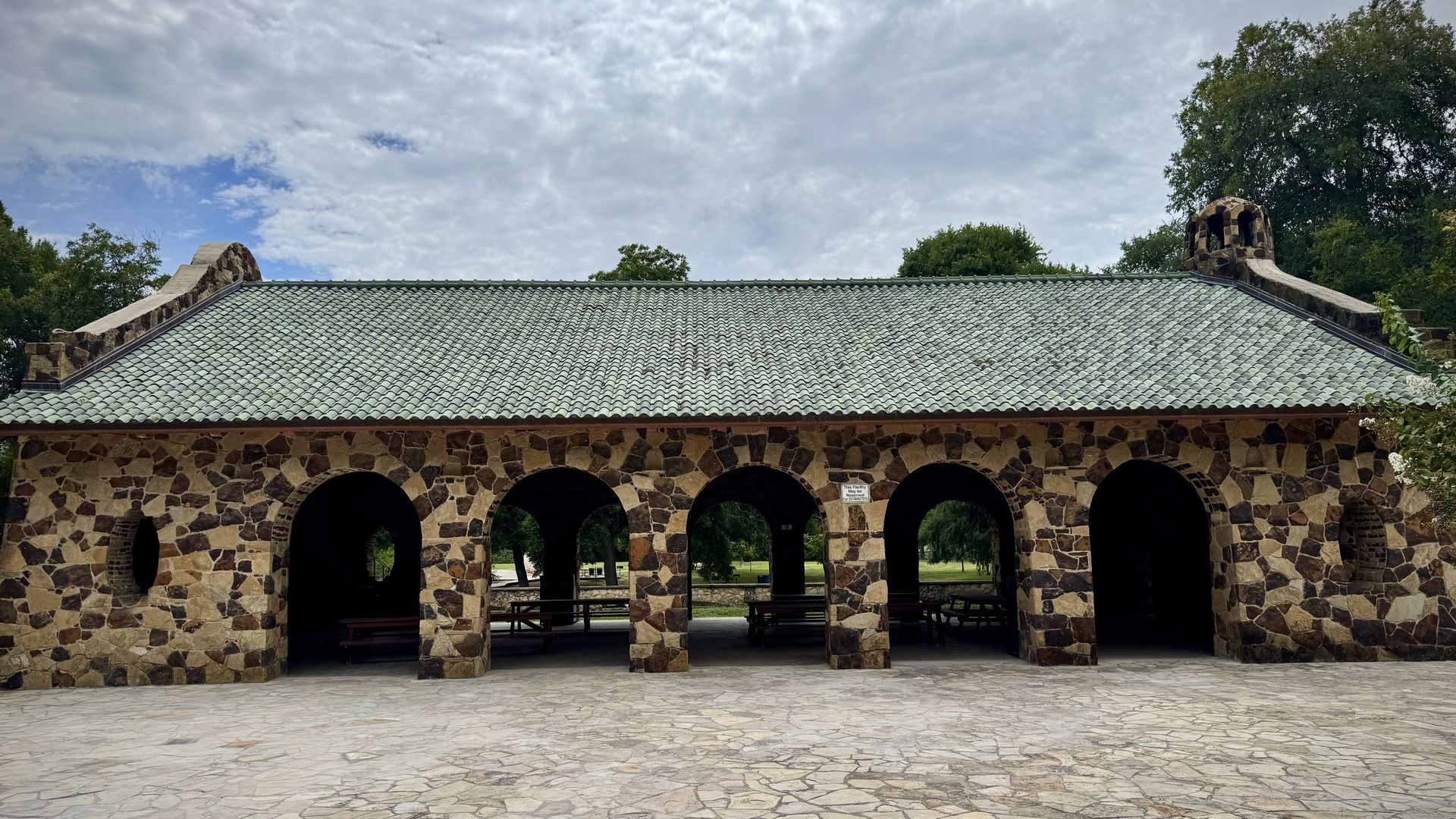Stone pavilion with five large archways, a green tiled roof and a stone patio in front, surrounded by trees under a cloudy sky.