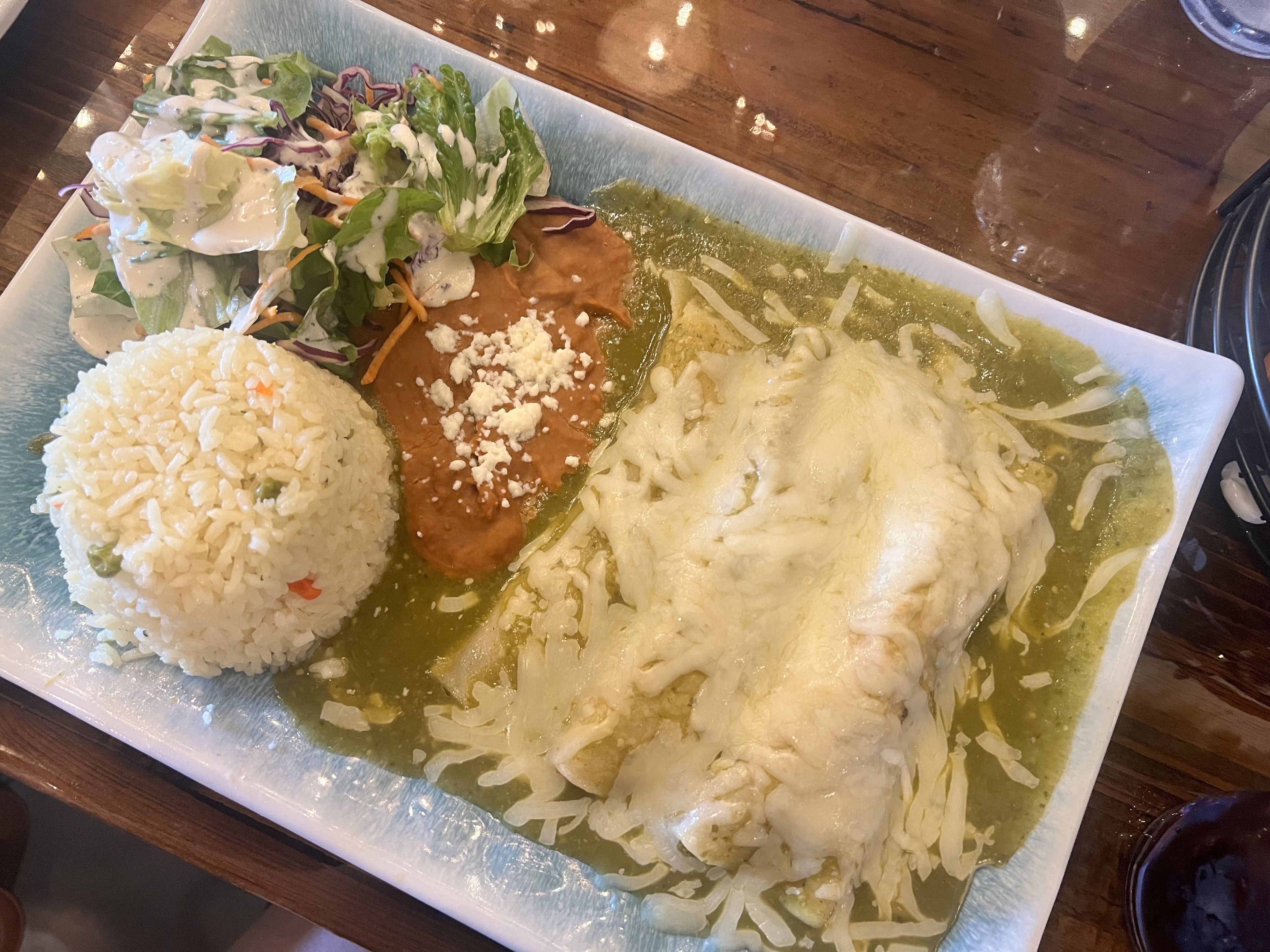 Rectangular plate with green salsa verde and melted cheese over chicken, a mound of rice, and a side salad with greens, purple cabbage, and carrots, all on a wooden table.