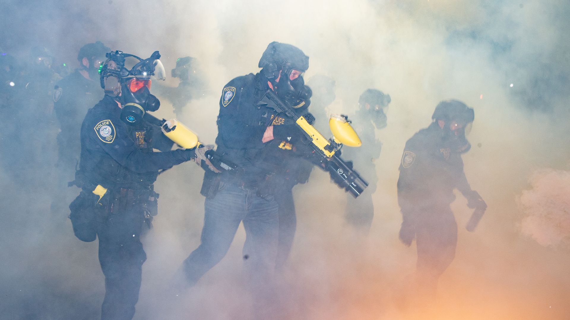 A squad of police in black tactical uniforms and gas masks advances through thick smoke, yellow-tipped rifles in hand, gear and patches visible, with an orange glow at the ground.