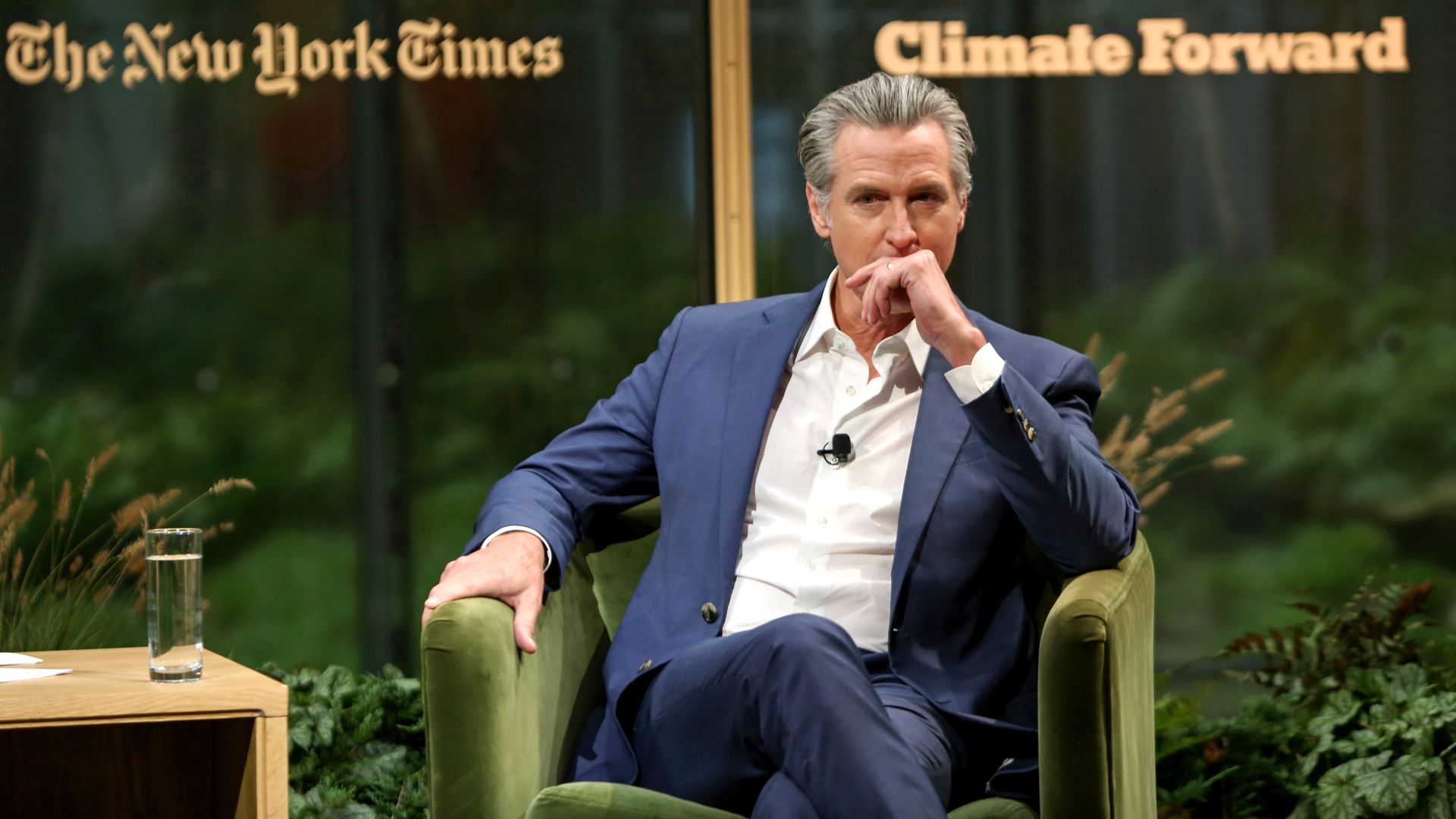 A man in a blue suit sits in a green armchair on a stage, hand to his mouth. Behind him, glass panels display "The New York Times" on the left and "Climate Forward" on the right, with greenery.