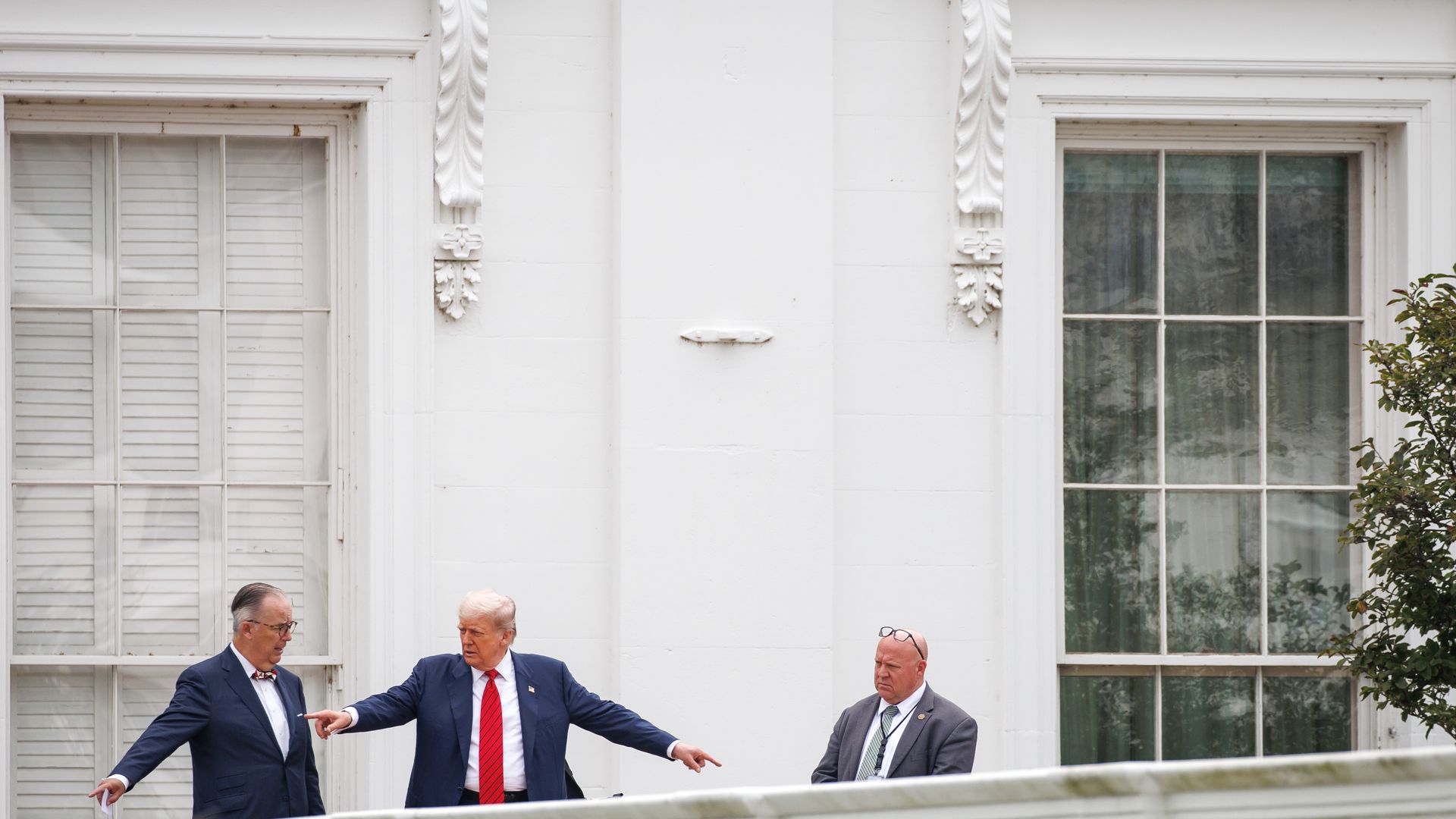 Three men in suits stand outside a white building with large windows and decorative trim. One man with a red tie gestures, another wears glasses on his head, and the third wears a bow tie.