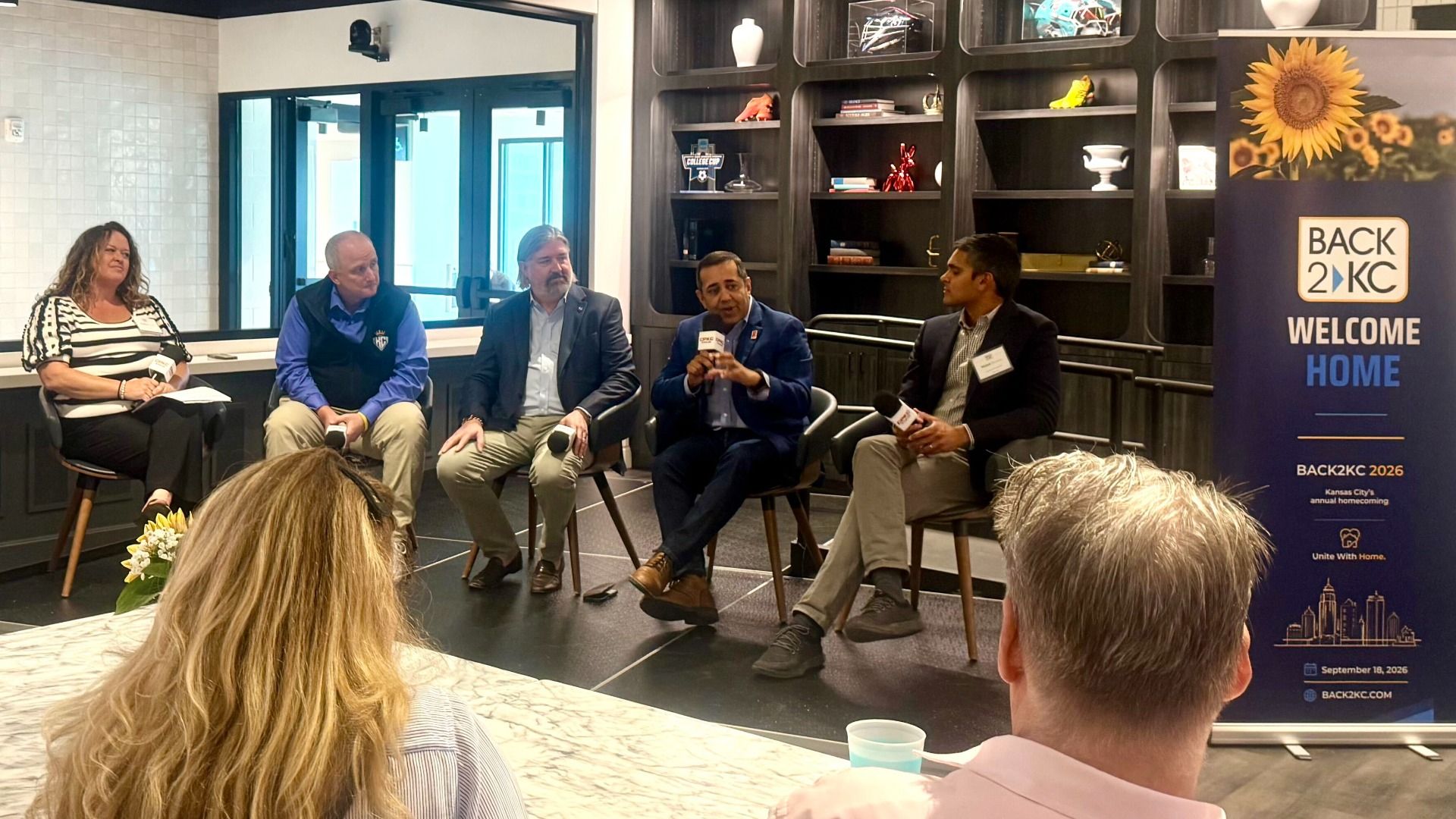 Panel discussion in a modern room with five speakers; a man in a blue suit speaks into a mic. A sunflower banner on the right reads "Back 2 KC Welcome Home"; audience visible in foreground.