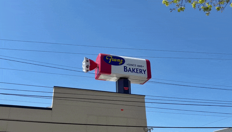 Animated sign shaped like a loaf of bread atop a building reads Franz Bakery, Since 1906, with the wrapper twisting as if unwrapping. Blue sky, power lines crossing, tree leaves in corner above the roof.