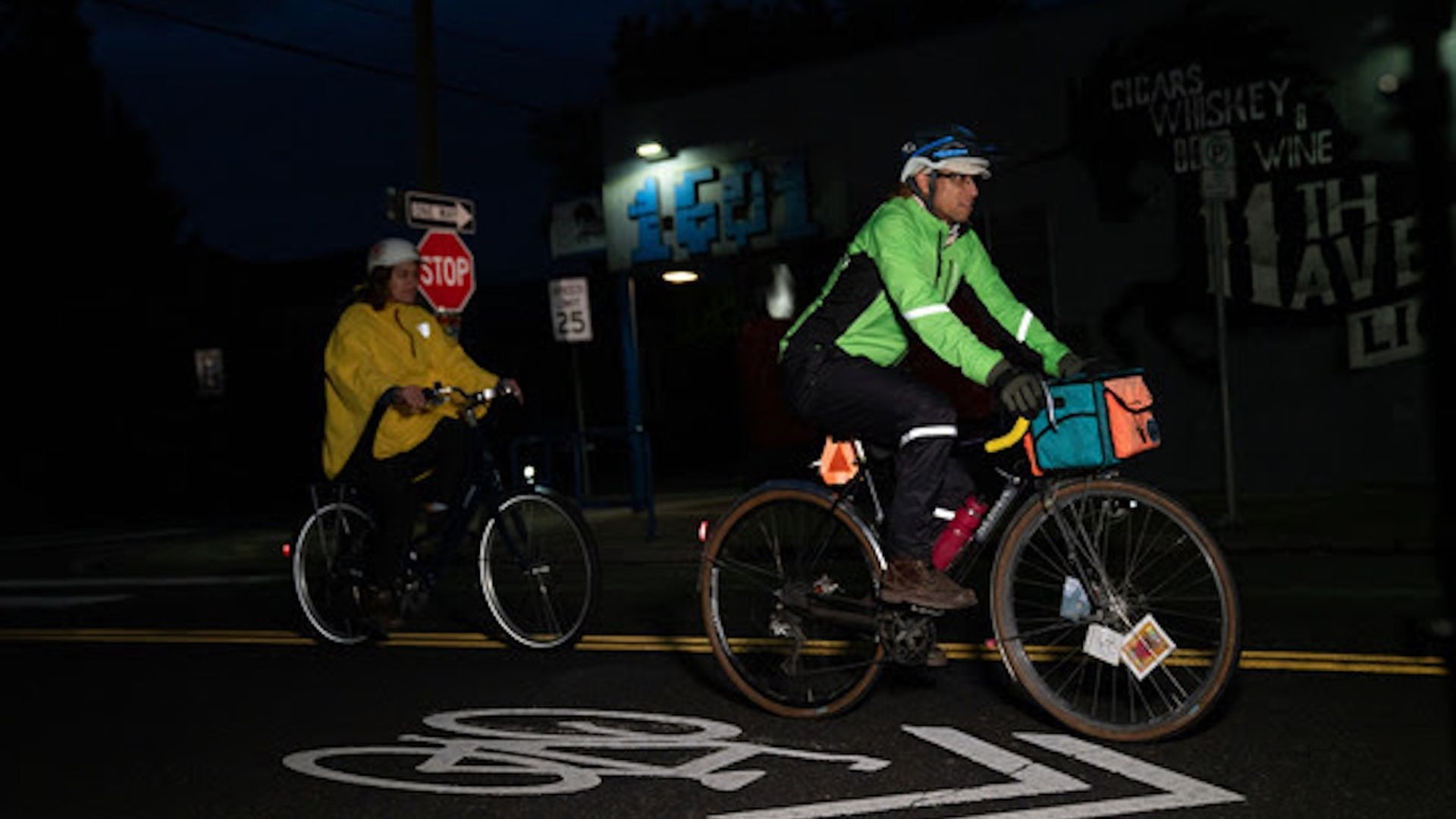 Two cyclists wearing helmets ride at night on a street with a painted bike lane symbol. One wears a bright green jacket, the other a yellow jacket, passing a stop sign and speed limit 25 sign.