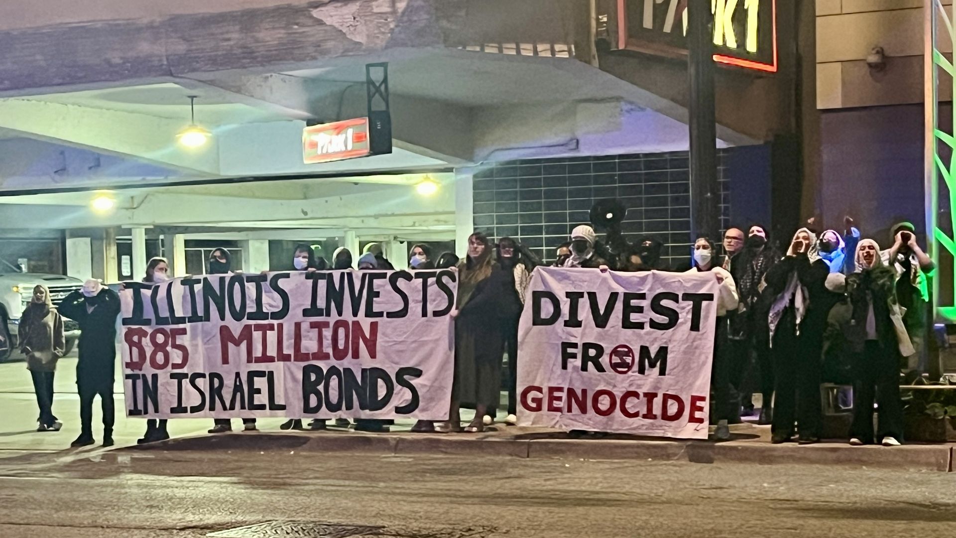 People standing in front of a parking garage holding signs that read Illinois Invests $85 million in Israel bonds and another that reads Divest from Genocide.