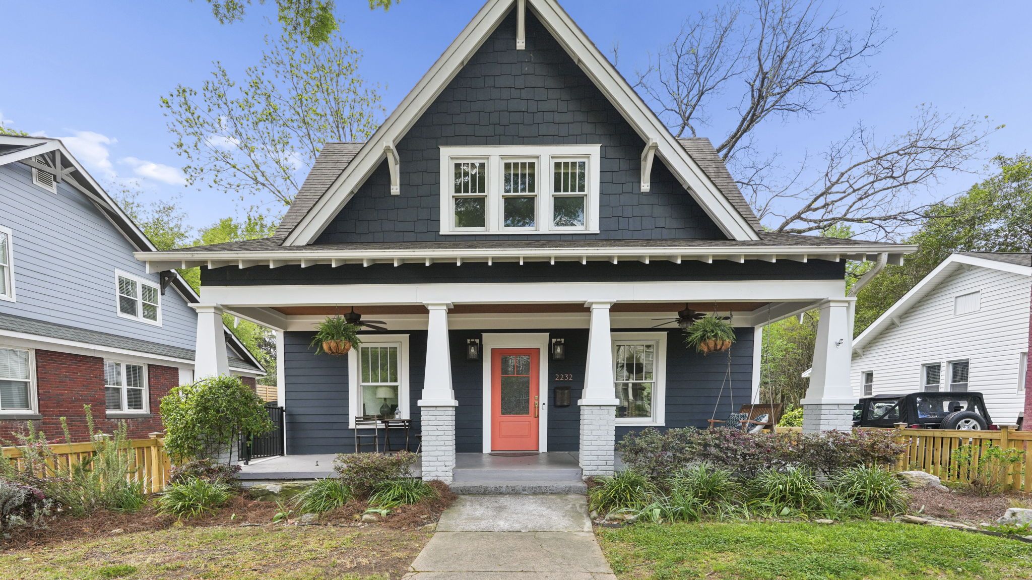 Charming blue craftsman-style house with a coral front door, white trim, and a wide porch. Hanging planters under the roofline; neighboring houses and trees line the street.