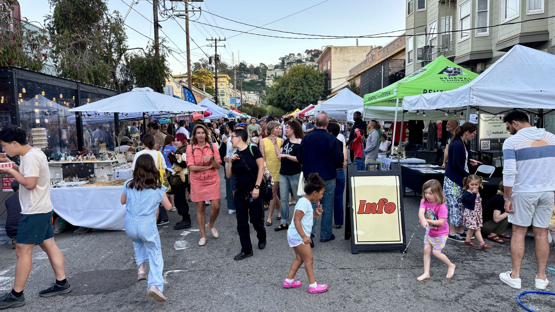 A bustling street market with white and colorful tents lining the street, crowds browsing stalls, people chatting, and children walking near buildings in the background.