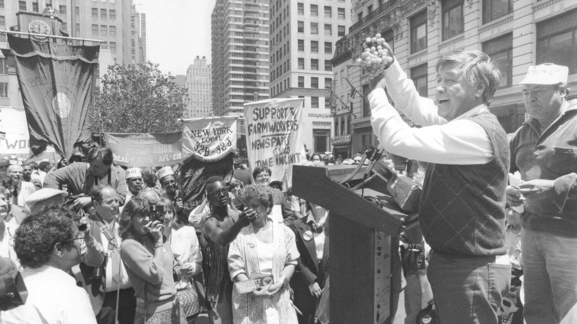 Black-and-white street rally: a man at a podium raises a bunch of grapes to a cheering crowd; banners read 'Support Farm Workers' with photographers and onlookers, while tall city buildings loom in the background.