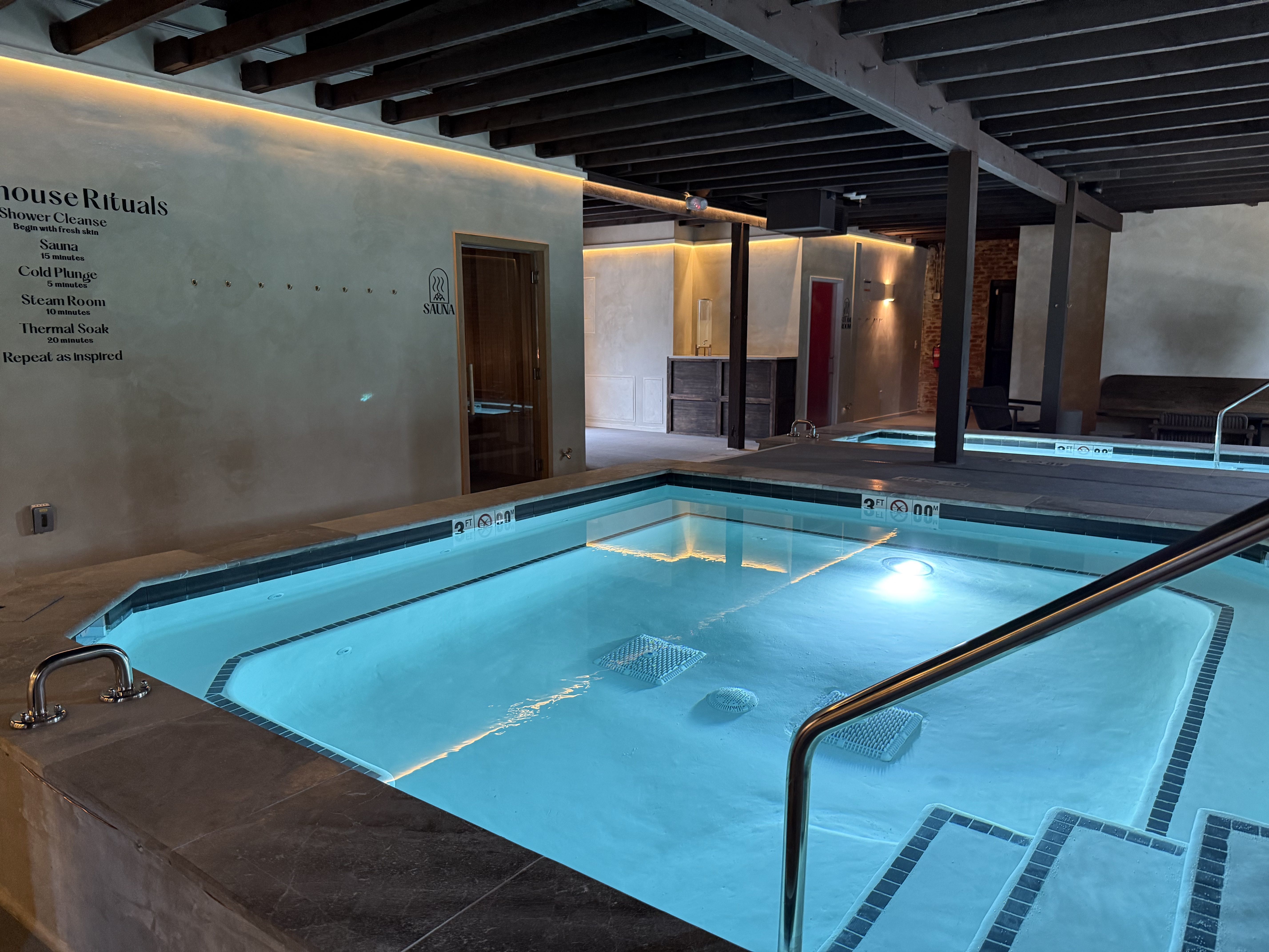 Indoor spa area with illuminated blue pools, a wooden-beamed ceiling, beige walls, a sauna door, and a wall listing "House Rituals" for shower cleanse and thermal soak steps.