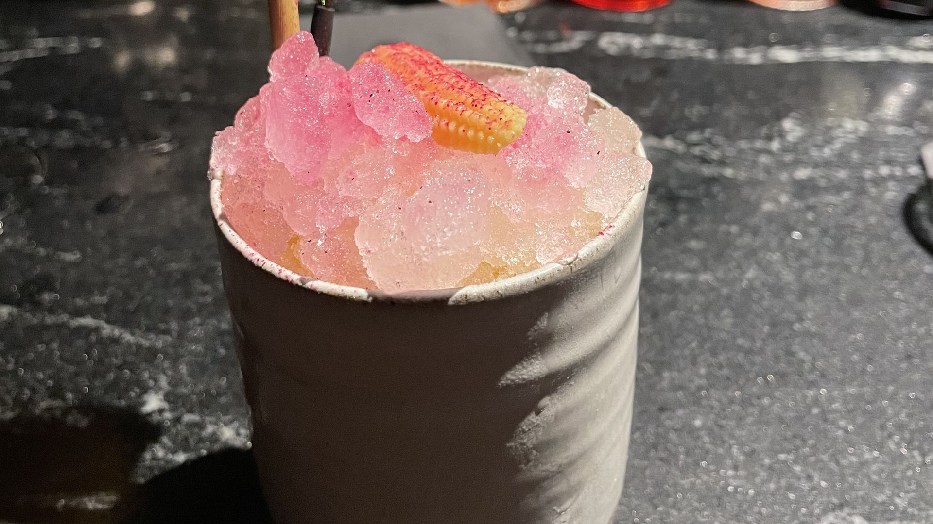A white mug showing a tequila cocktail topped with ice and pink dragonfruit powder on top on a bar counter.
