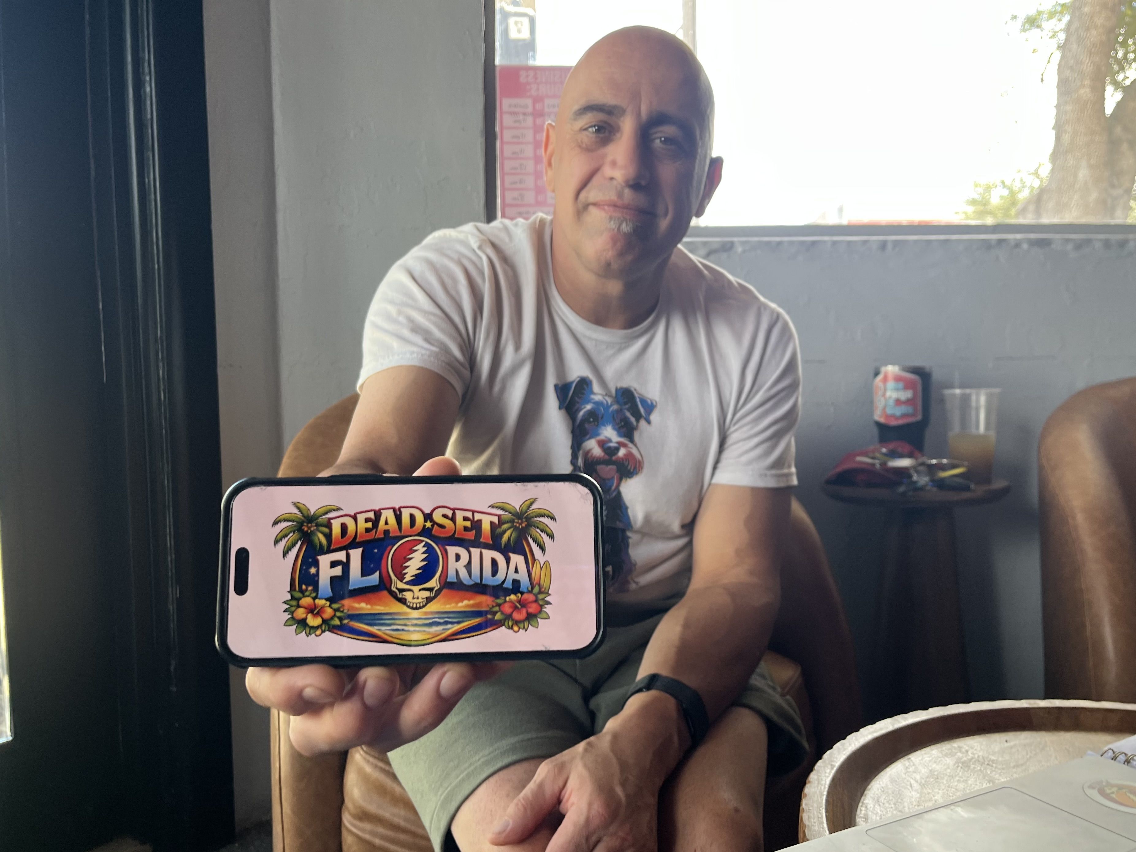 Smiling bald man in a white T-shirt sits indoors, holding a phone showing a tropical poster that reads "DEAD-SET FLORIDA" with palm trees and a skull; window with a tree behind.
