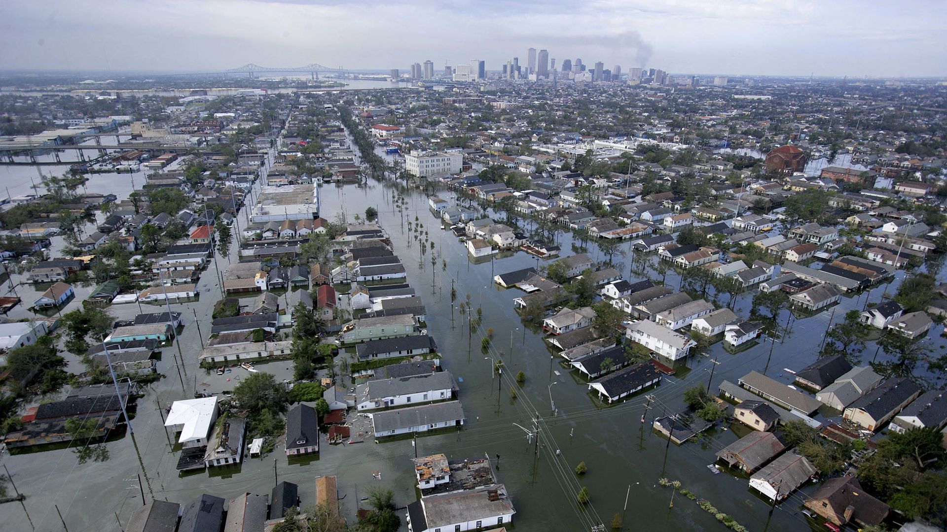 Aerial view of a city heavily flooded with water covering streets and reaching house rooftops, with a distant city skyline under a cloudy sky.