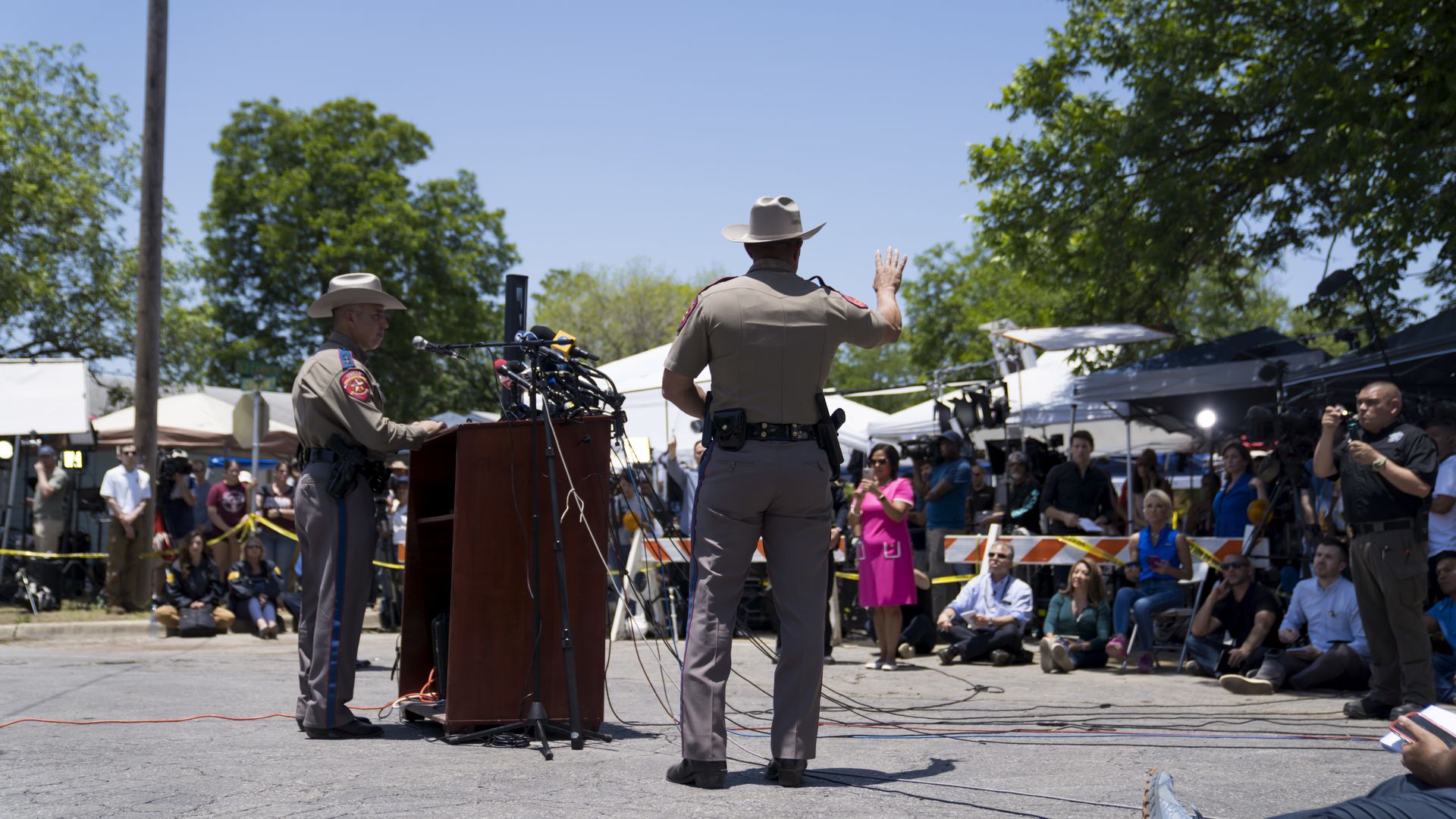 A Texas Department of Public Safety office waves his hand at reporters at a news conference in Uvalde. Another officer stands in front of a podium with many microphones on it. 