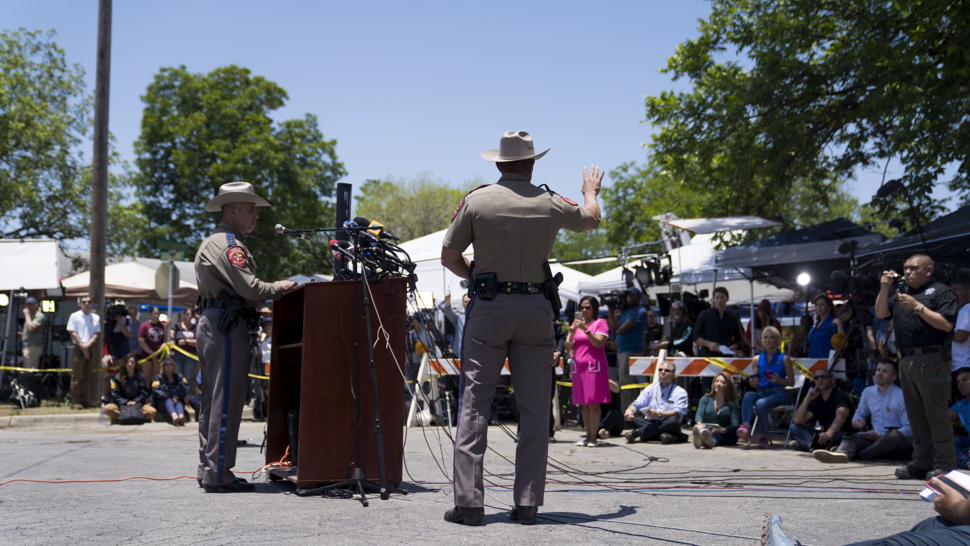 A Texas Department of Public Safety office waves his hand at reporters at a news conference in Uvalde. Another officer stands in front of a podium with many microphones on it.